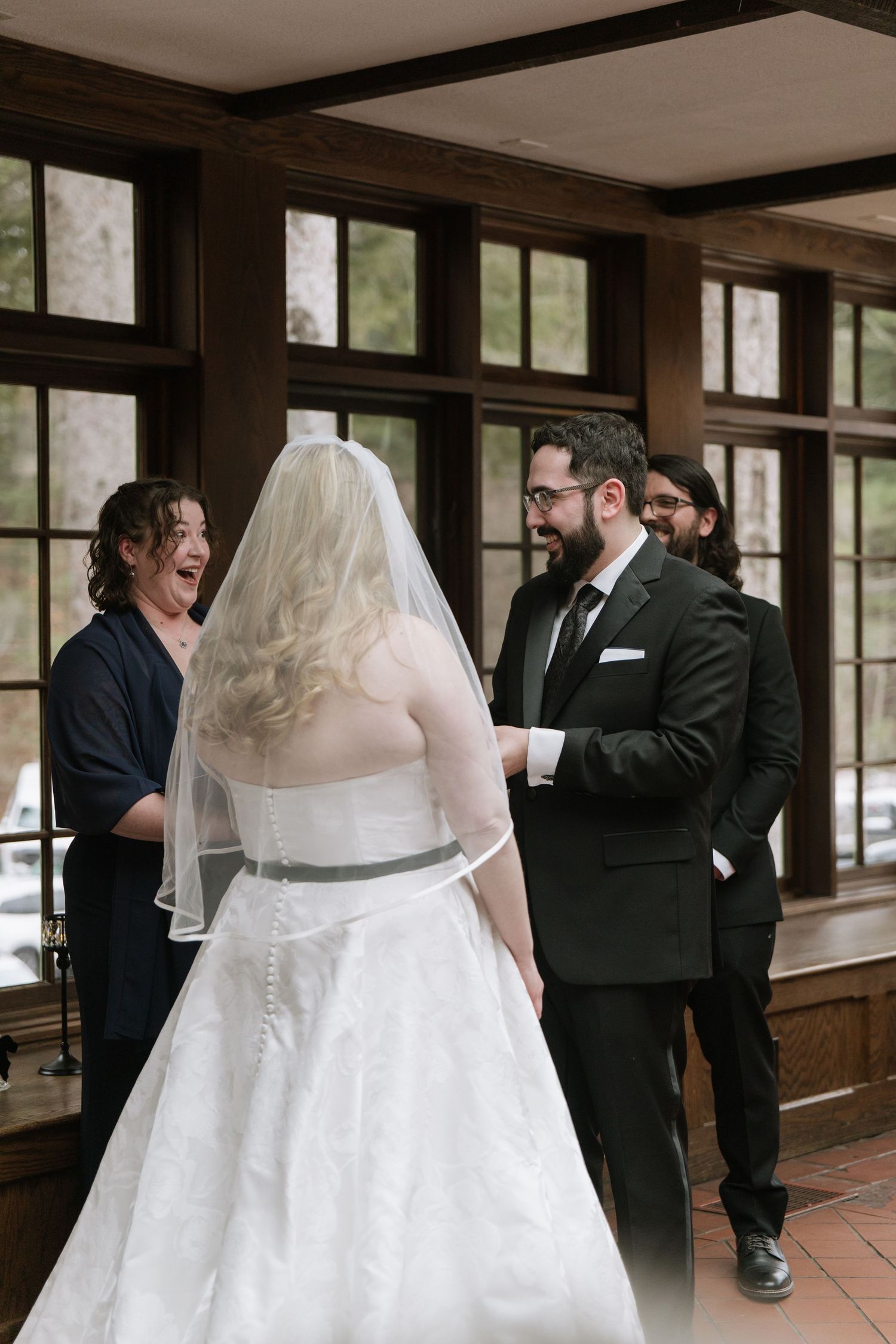 Bride in white gown and veil shares a joyful first look moment with groom in black suit at indoor wedding venue.