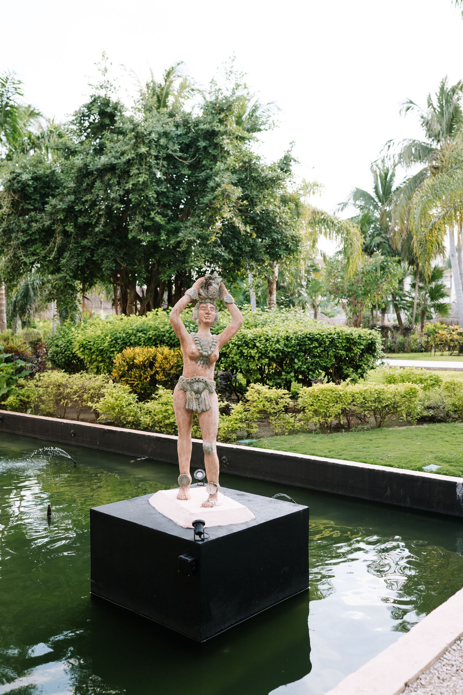 Outdoor water feature with dark reflecting pool surrounded by lush tropical garden and palm trees.