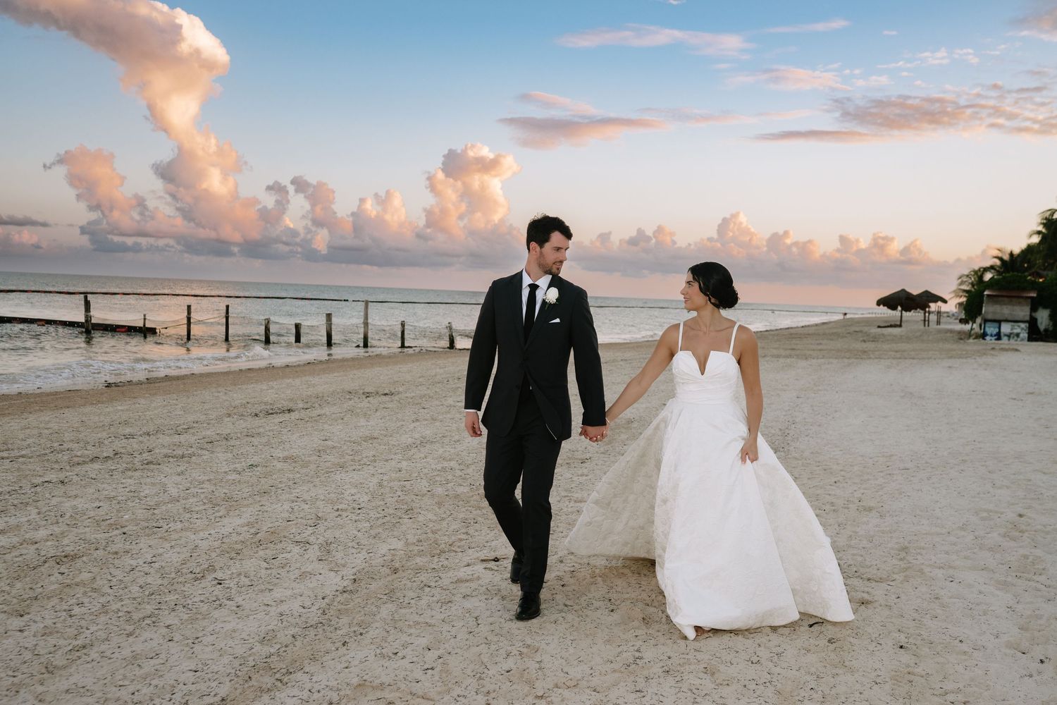 A couple walks along a sandy beach at sunset with pink clouds in the sky as their white dress and black suit flow in the wind.