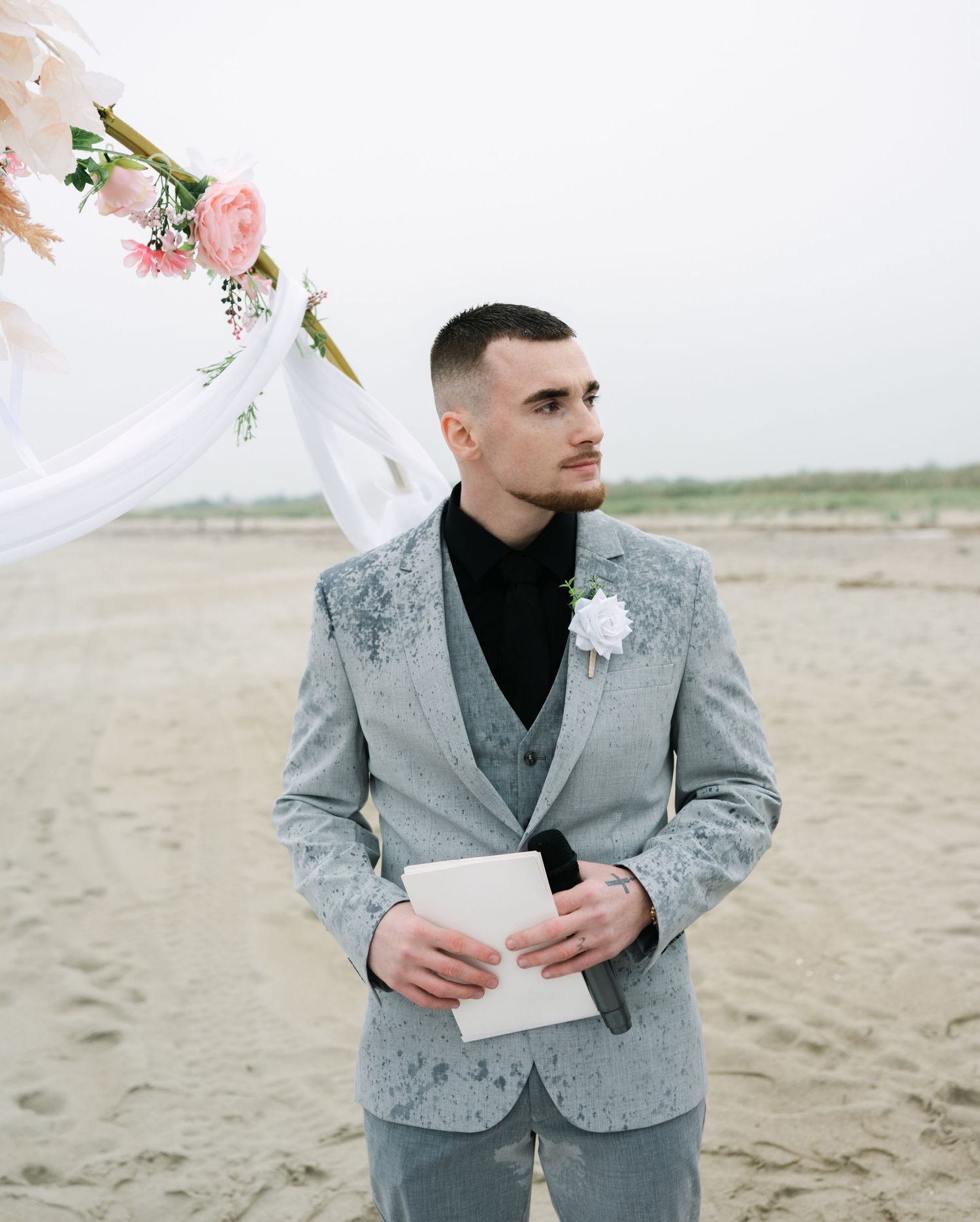 A person in a grey suit jacket and black turtleneck reads from paper while standing on a beach.