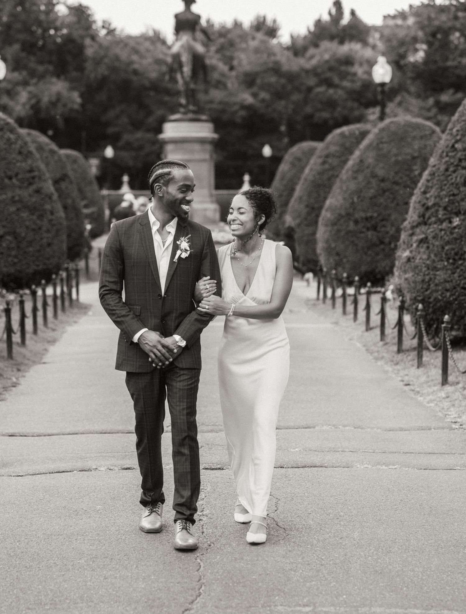 Black and white photo of a couple in wedding attire walking together in a formal garden lined with sculpted hedges.