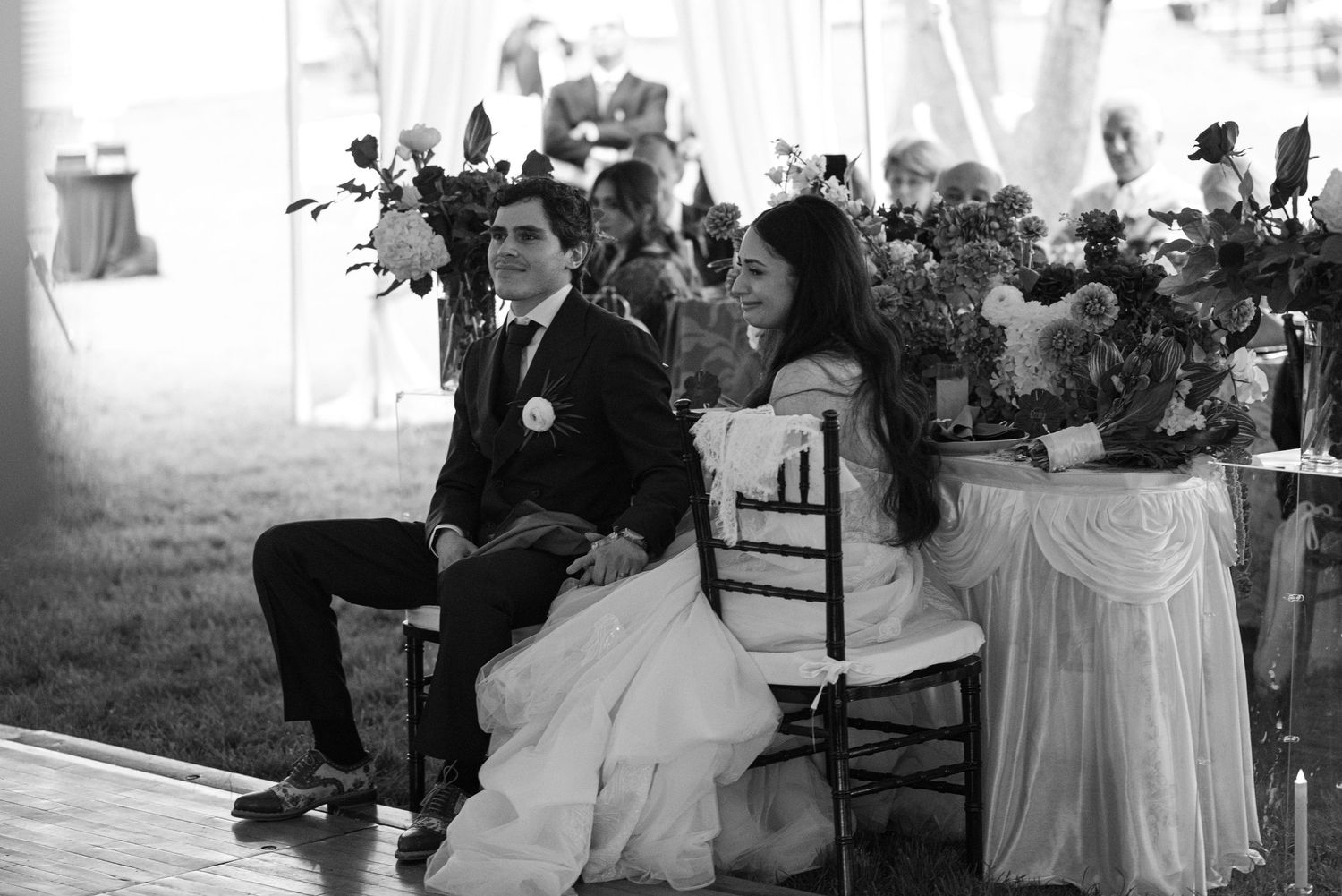Bride and groom seated together at sweetheart table during wedding reception under a white tent.