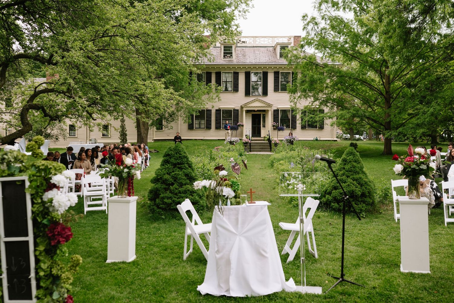 Elegant outdoor wedding ceremony setup on green lawn before historic colonial mansion with white chairs and floral arrangements.