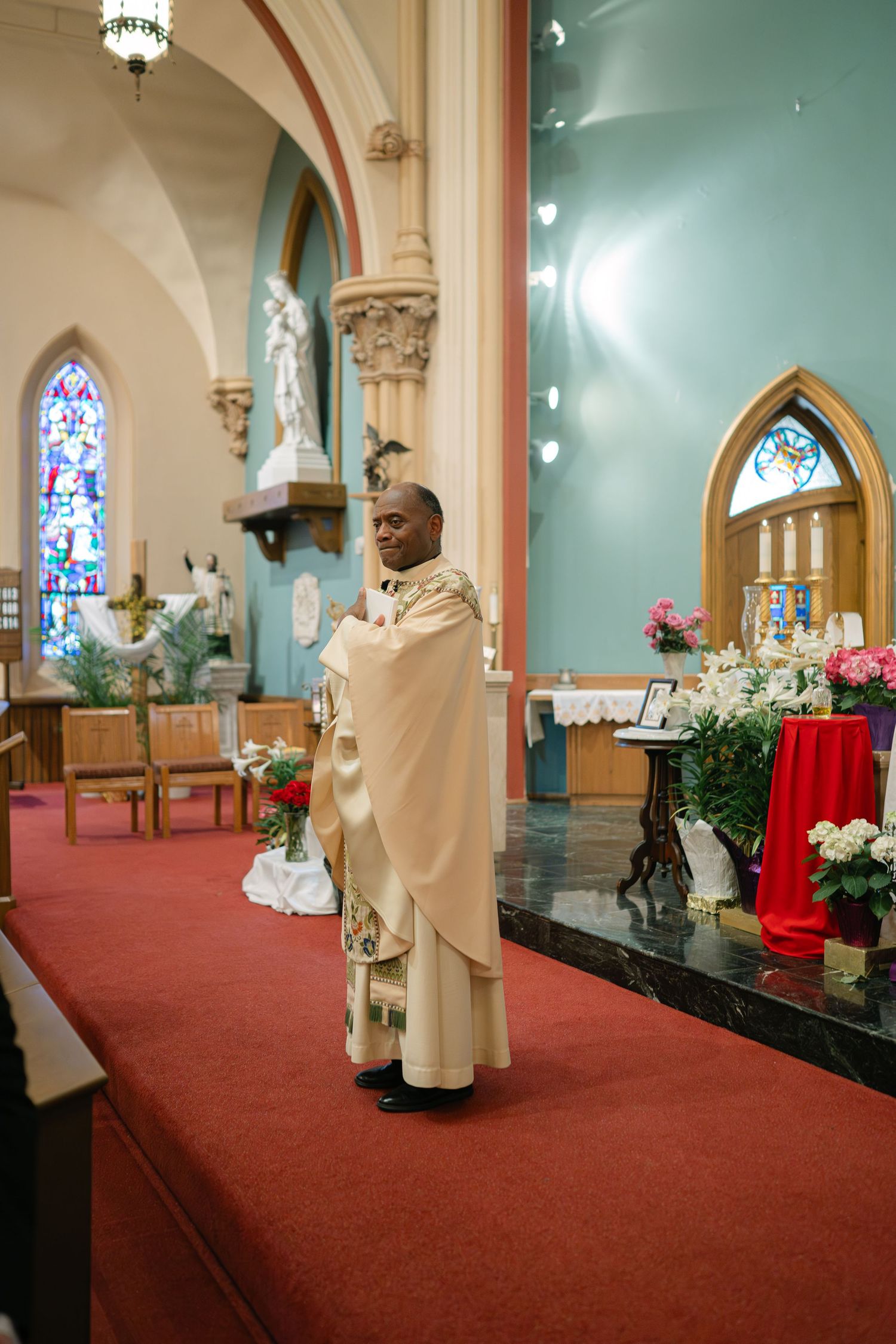Religious ceremony taking place in a church sanctuary with stained glass windows, red carpet, and floral arrangements.