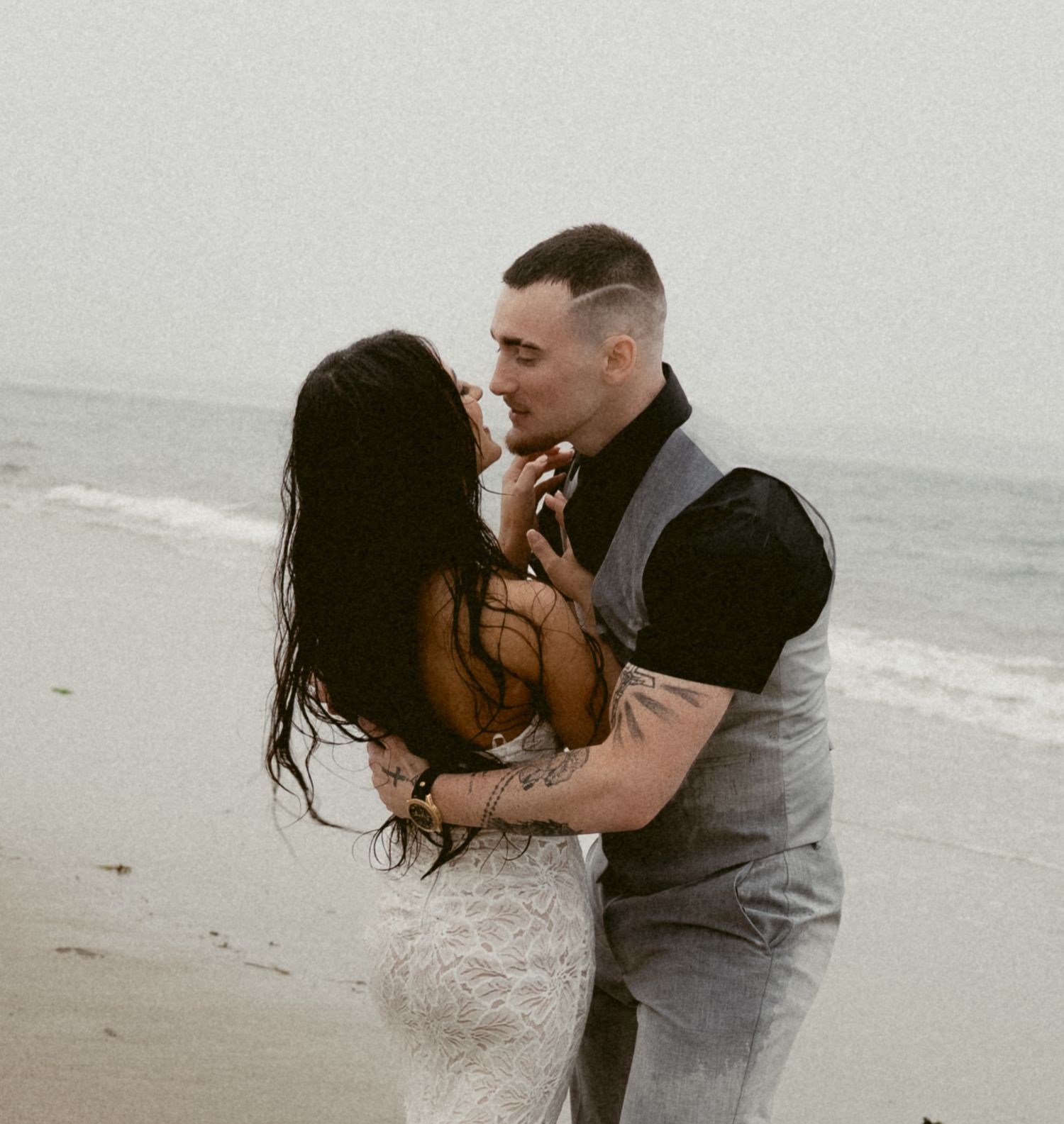 A romantic couple embraces on a misty beach shore as waves crash behind them in black and white tones.