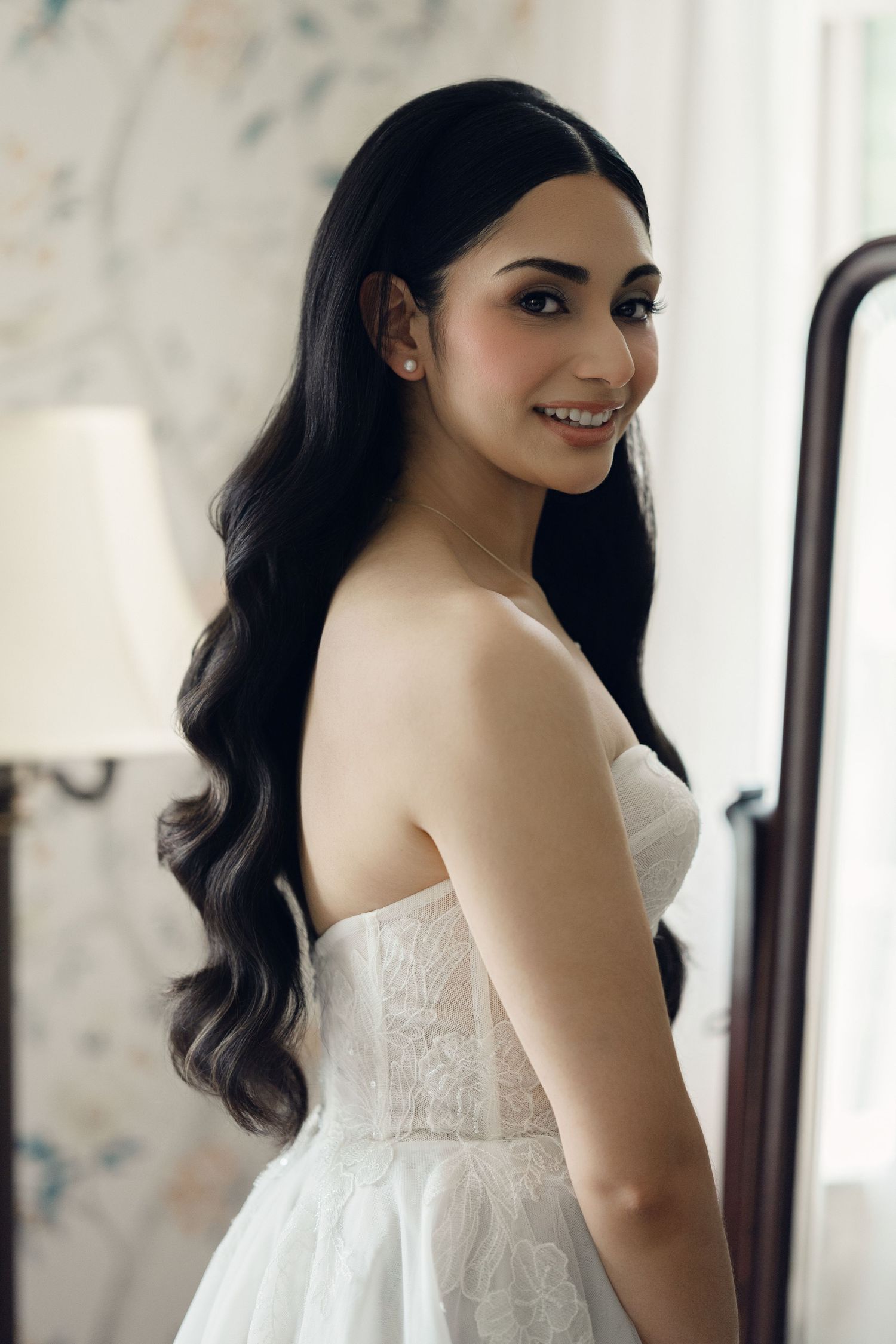 A smiling bride in a strapless white lace wedding gown with long wavy dark hair poses elegantly in a bright room.