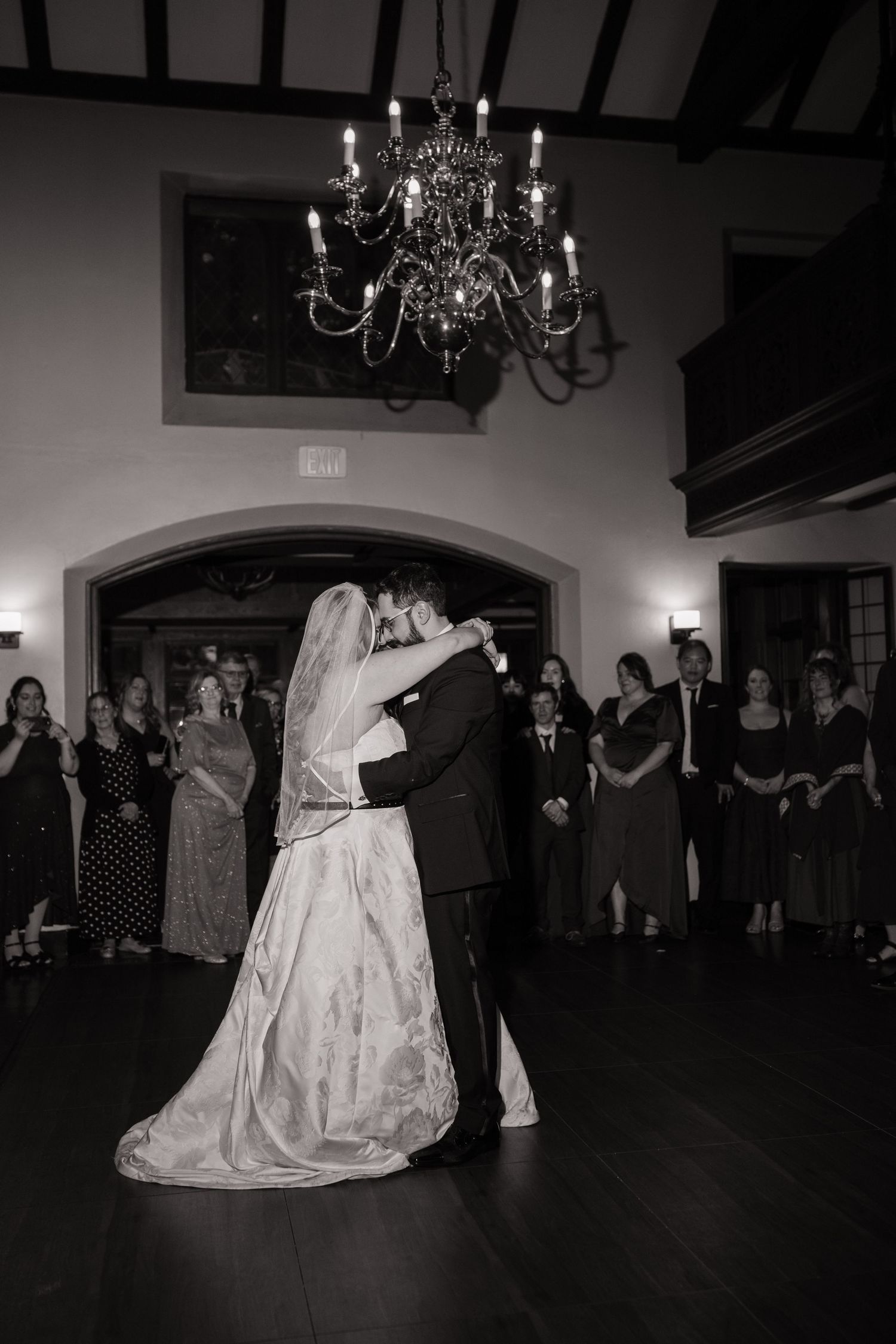 Black and white photo of bride and groom sharing first dance under chandelier with guests watching at elegant wedding reception.