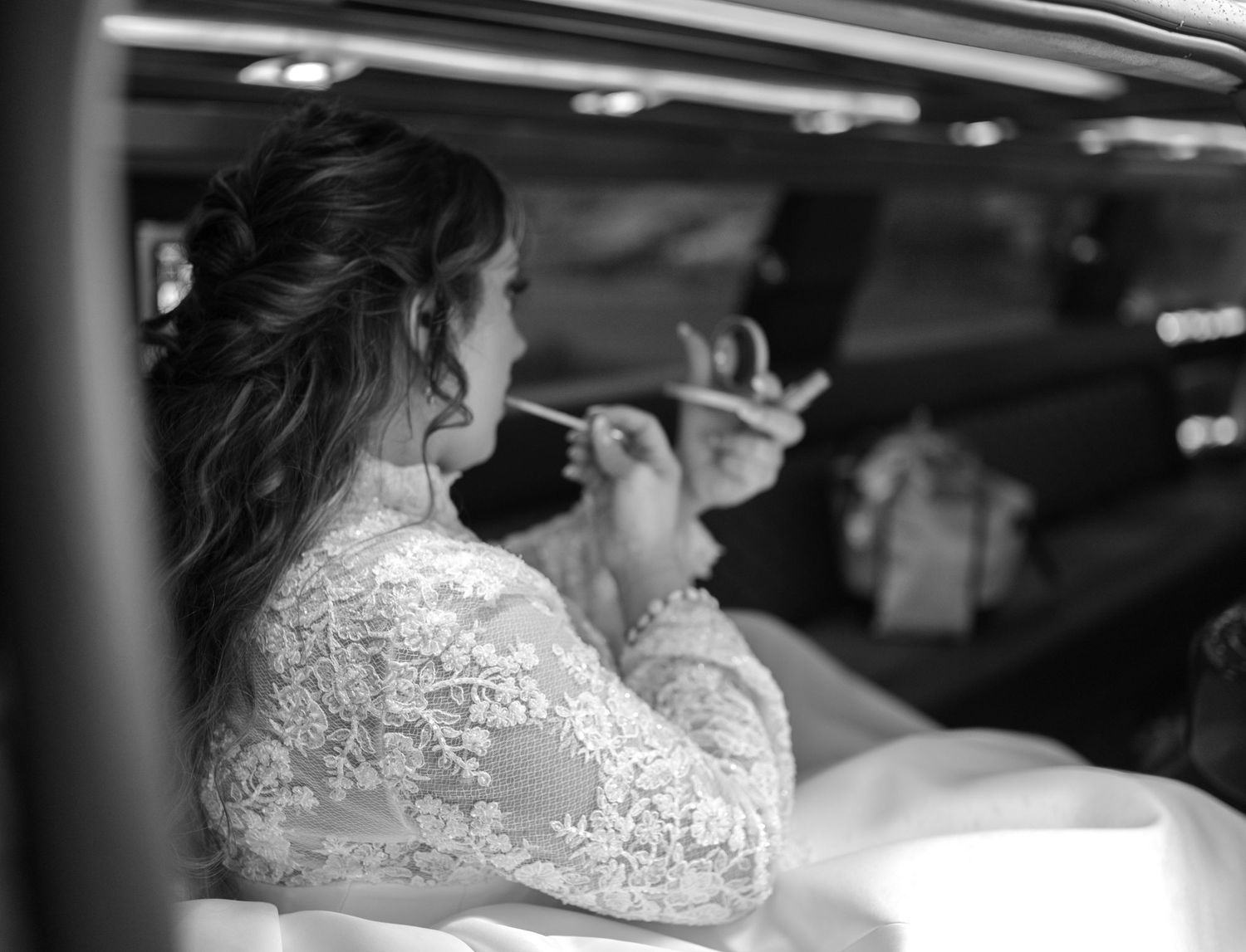 A black and white photo shows someone in a lace wedding dress touching up their makeup in a car.