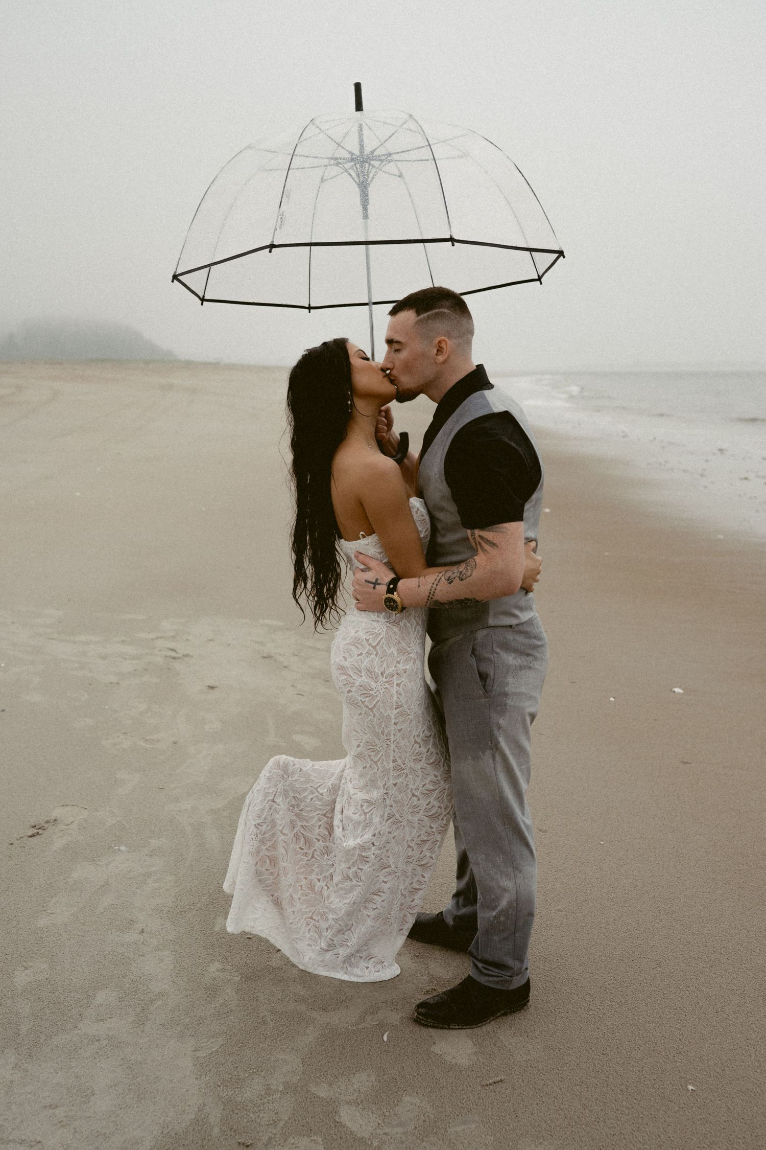 A romantic couple shares a kiss under a clear umbrella on a misty beach, with the bride wearing a lace wedding dress.