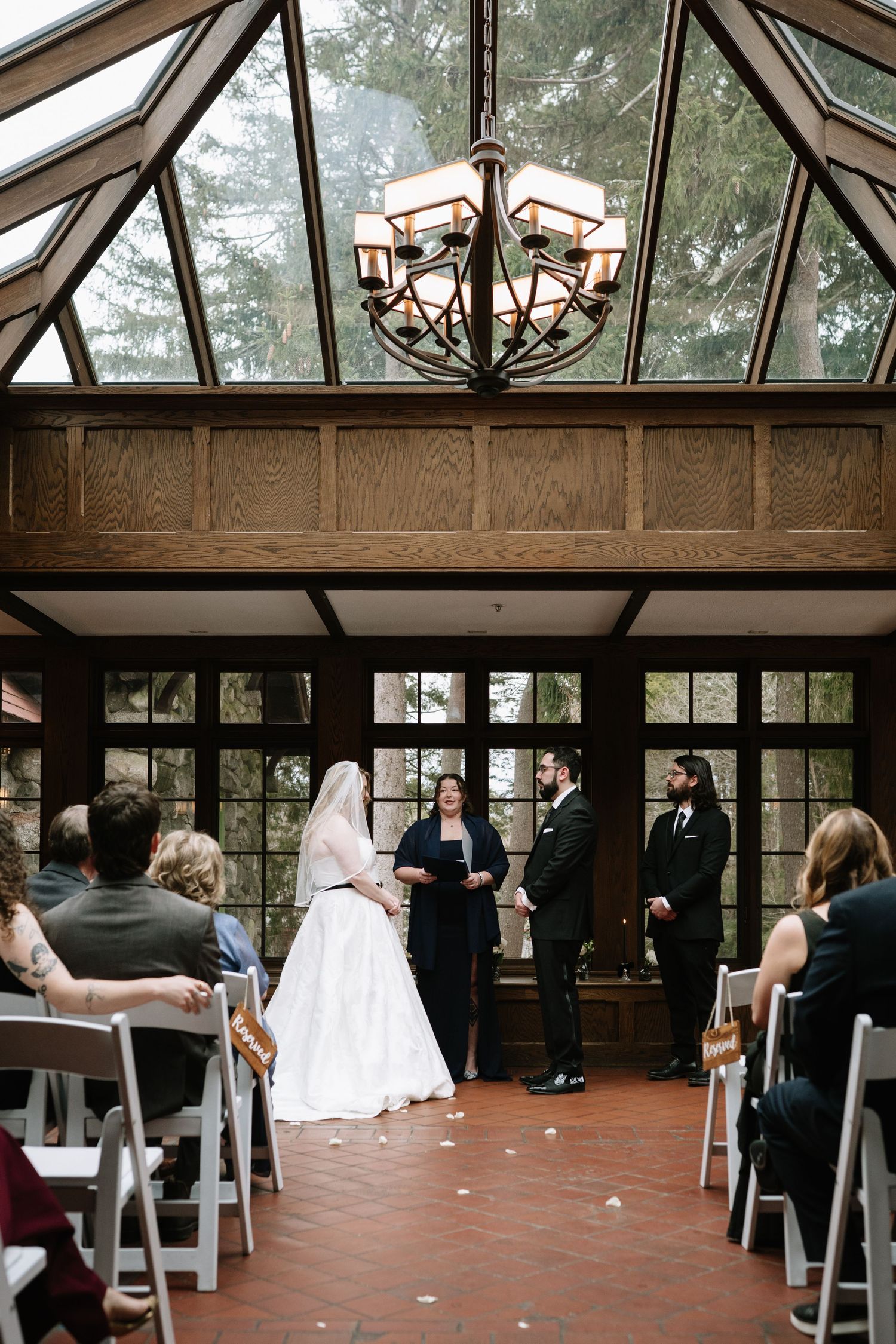 Wedding ceremony in elegant glass-ceiling chapel with chandelier, bride in white gown facing officiant and groom before seated guests.