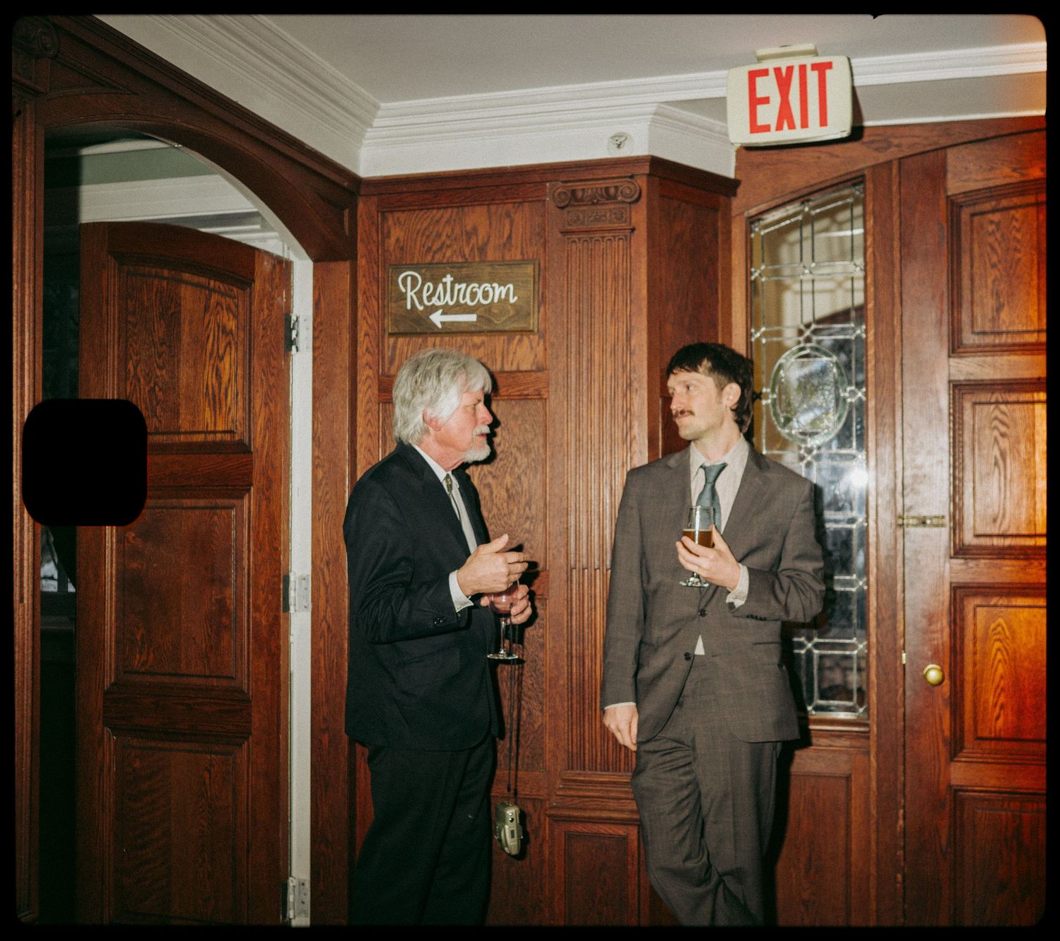 Two men in suits converse near restroom doors at a formal indoor event with wood-paneled walls.