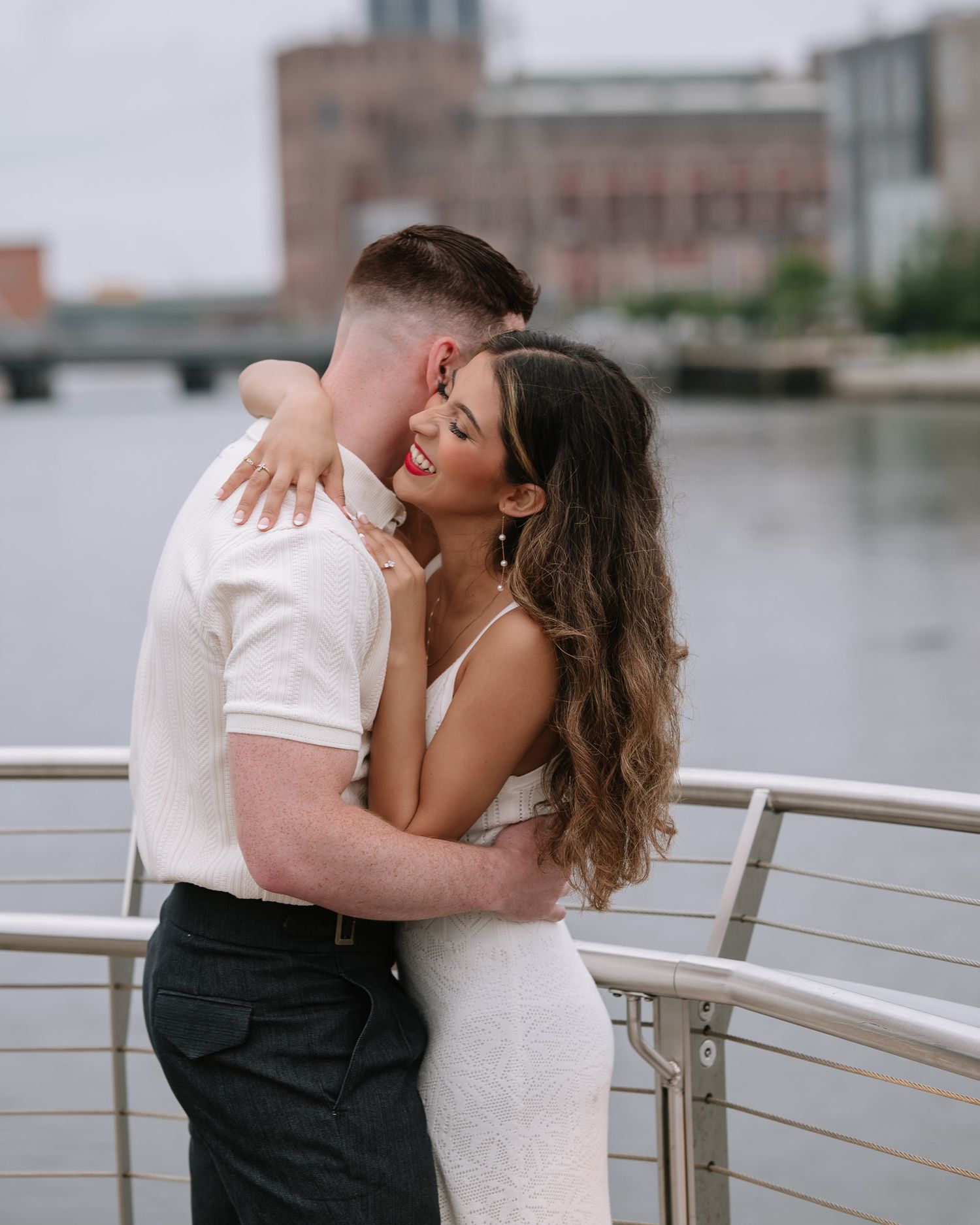 Couple in white and black attire sharing romantic moment by waterfront railing.