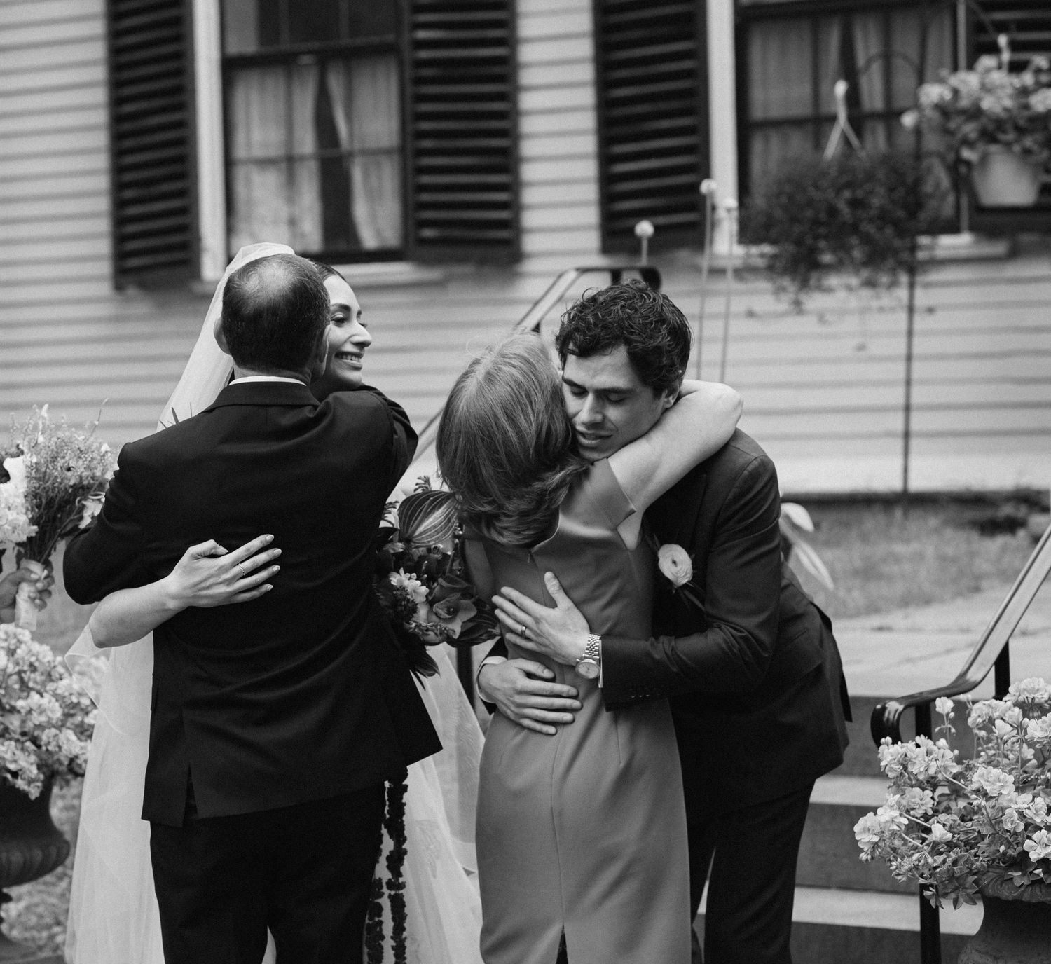 Black and white wedding photo of couples embracing and hugging outdoors near a house.