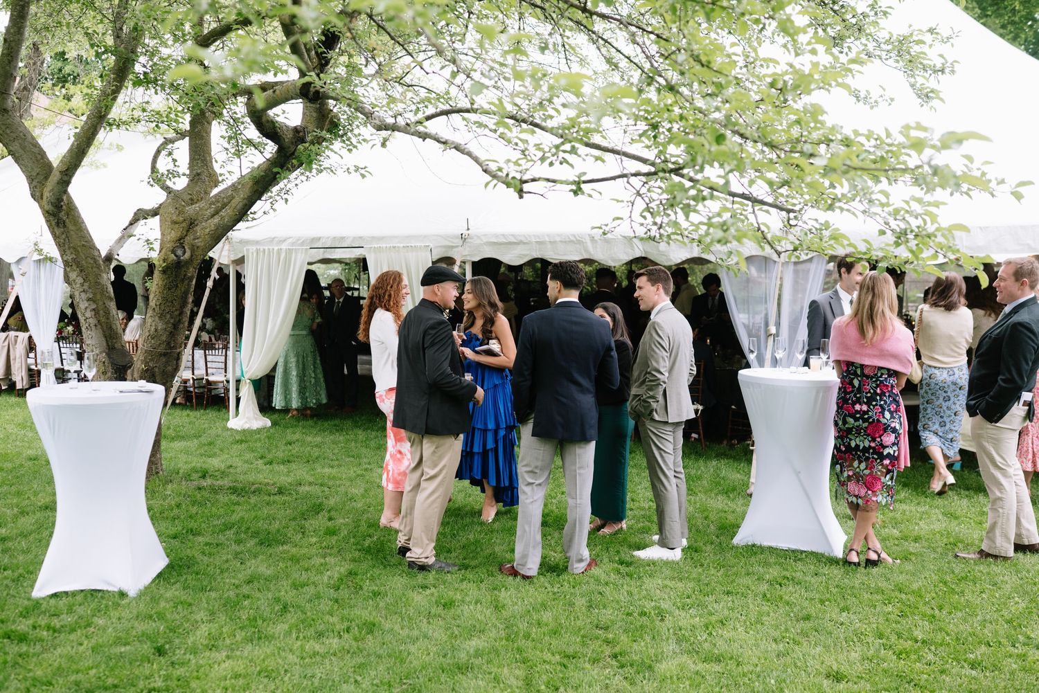 Elegant outdoor wedding cocktail hour on green lawn with white draped tent, high-top tables, and guests mingling under blooming trees.