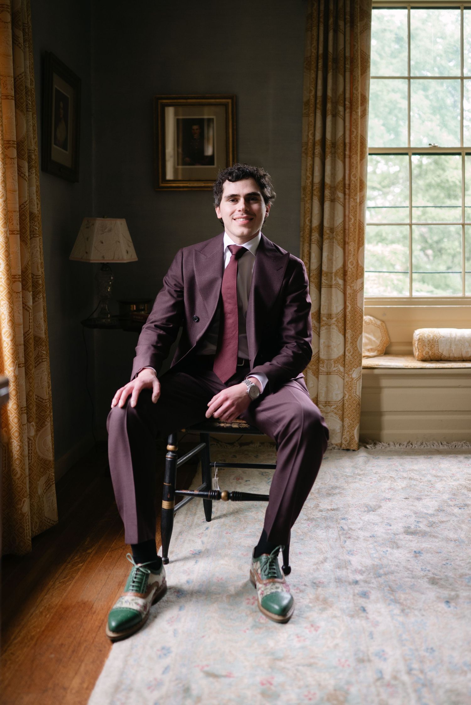 Young man in burgundy suit with green dress shoes seated by window in elegant room.