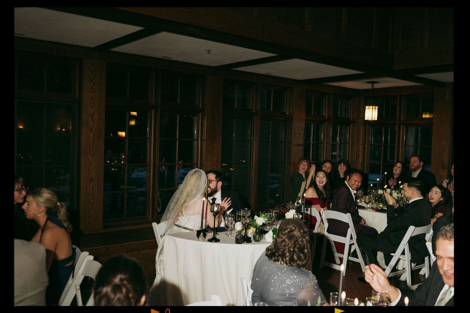 Bride and groom seated at wedding reception head table with guests in elegant wood-paneled venue.