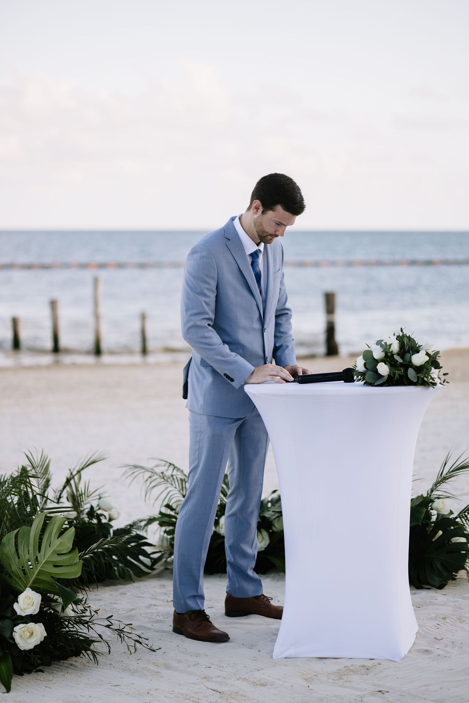 Person in light blue suit writing on paper at beachside setting with tropical greenery and ocean view.
