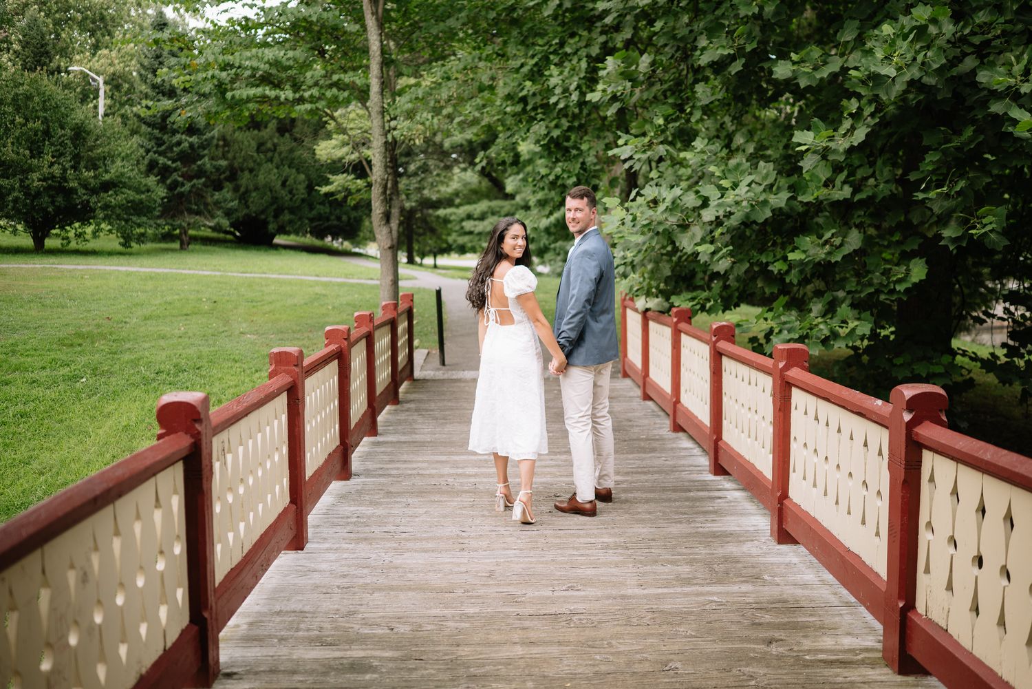 A couple walks hand in hand across a red-railed bridge surrounded by lush greenery.