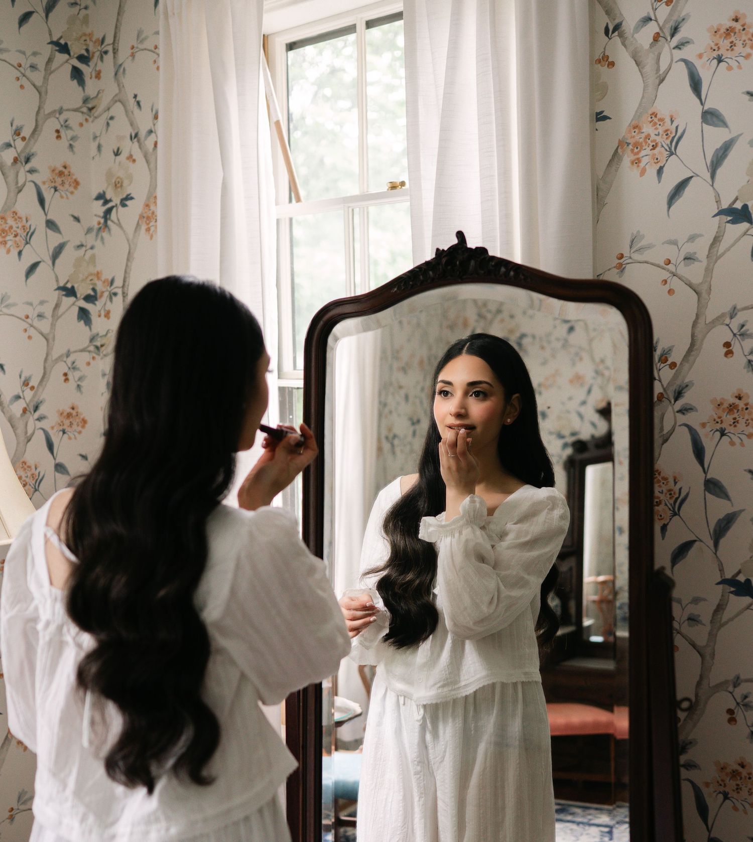 Woman in white dress applying makeup at ornate mirror in floral wallpaper bedroom.