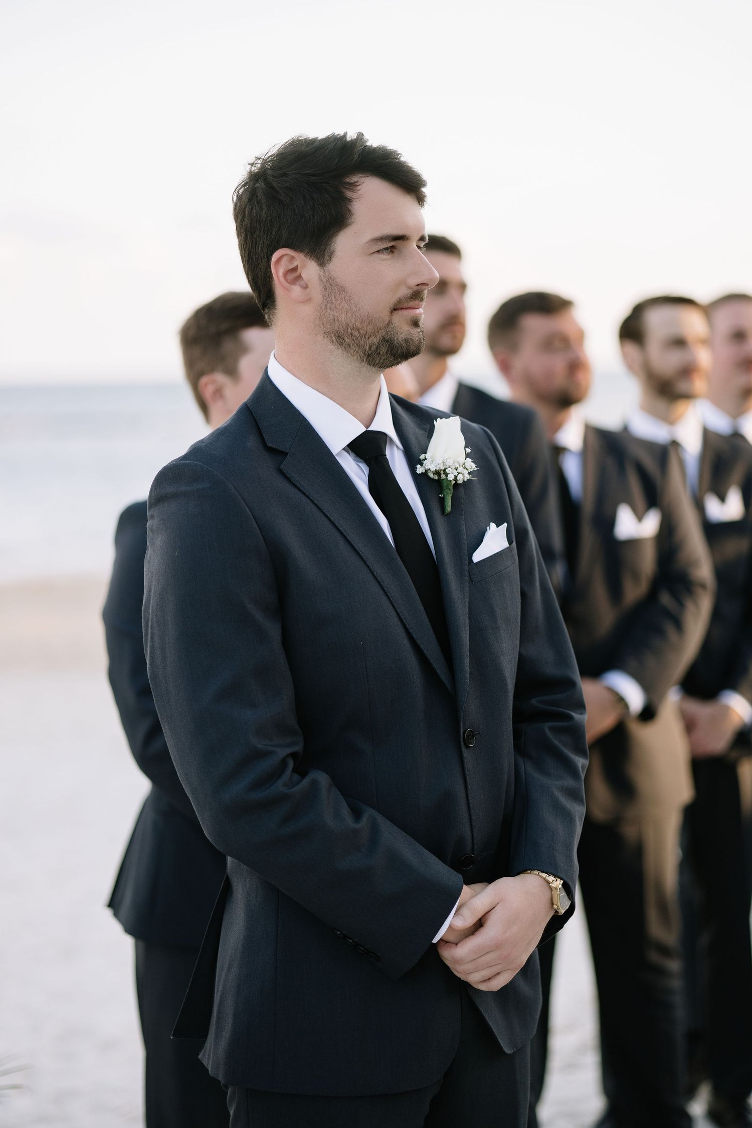 Wedding party stands at beachside ceremony wearing formal black suits and boutonnieres against ocean backdrop.