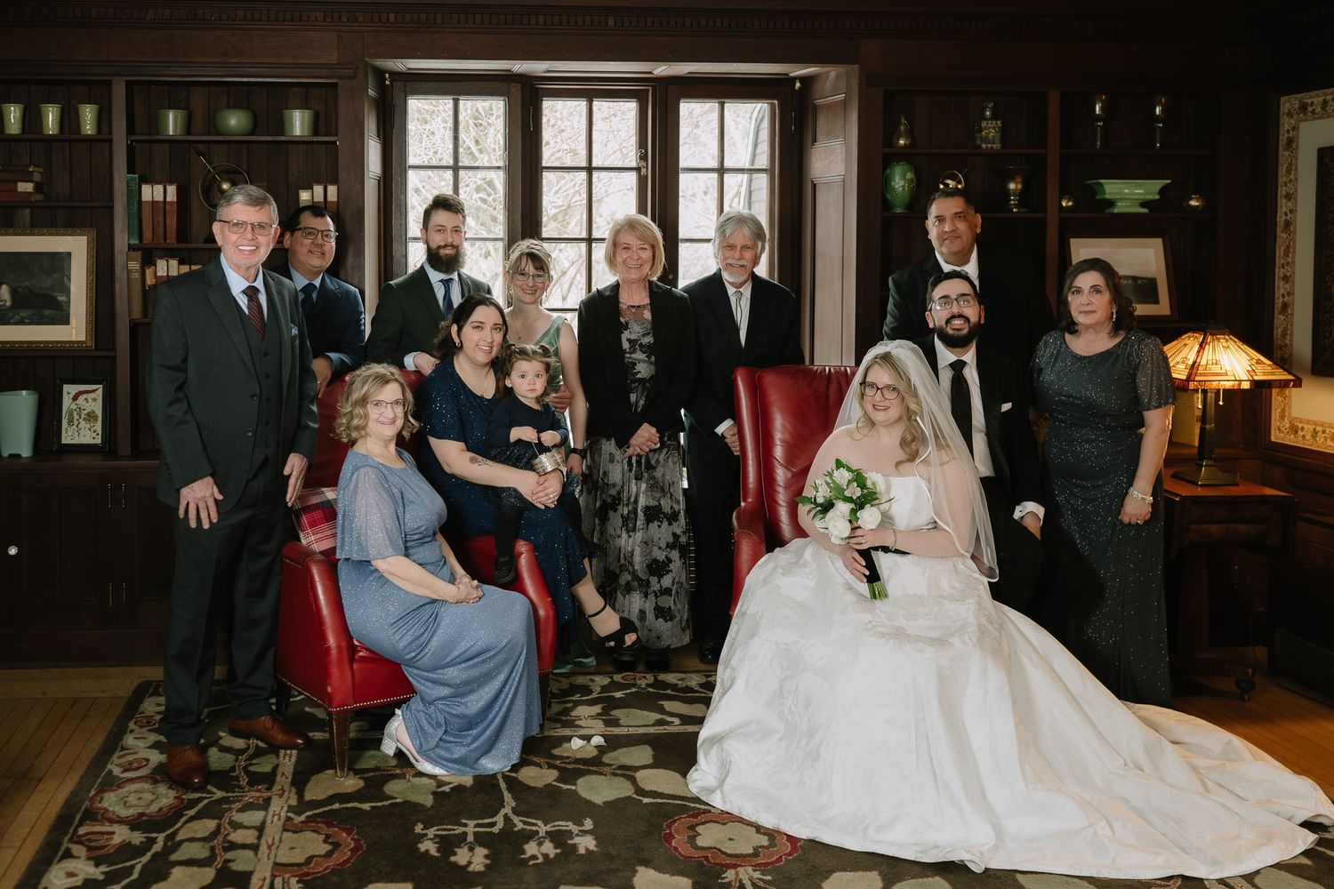 Wedding party family portrait in elegant wood-paneled room, bride in white gown seated with bouquet surrounded by formal guests.