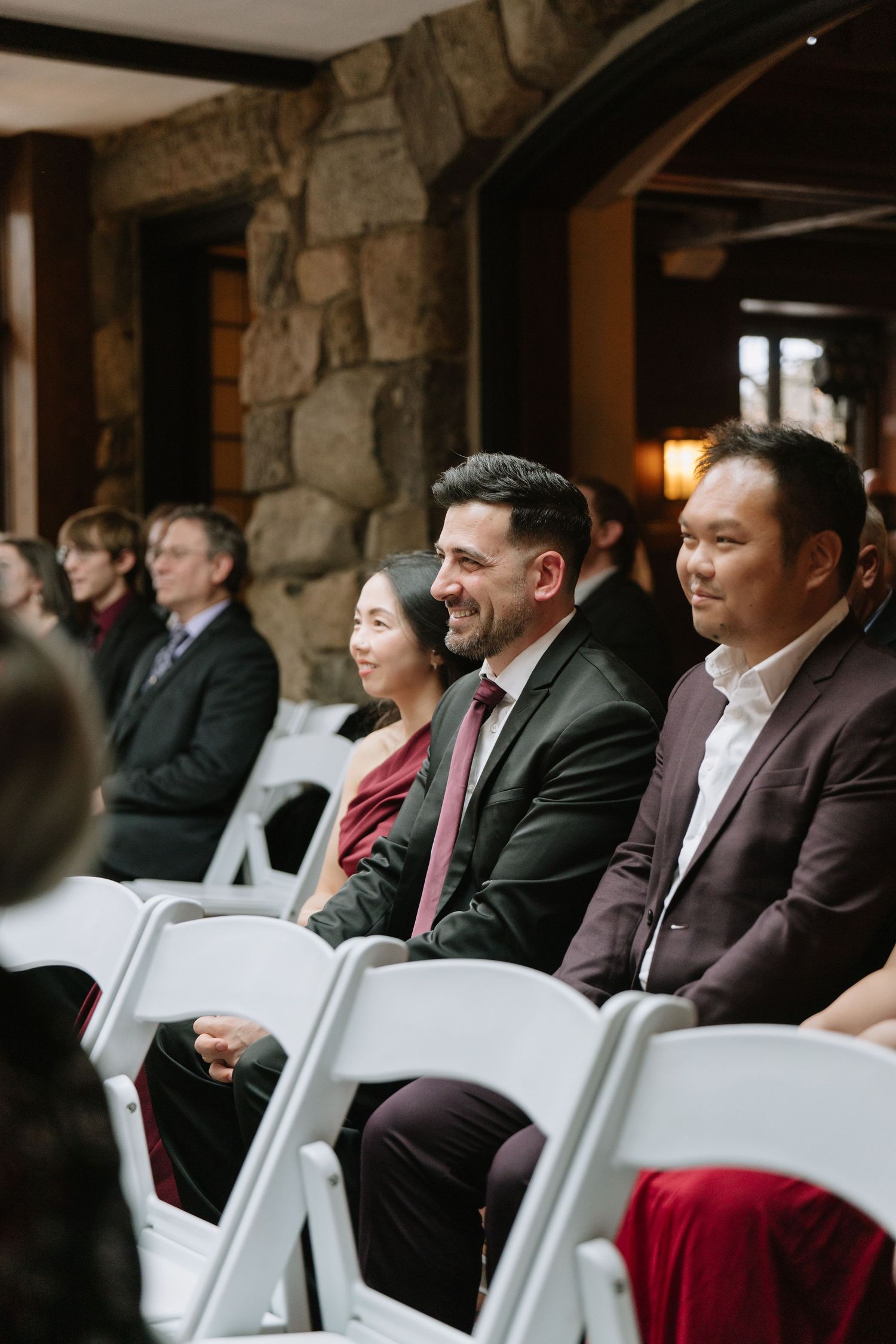 Wedding guests seated in white chairs smile during an indoor ceremony in a rustic stone venue.