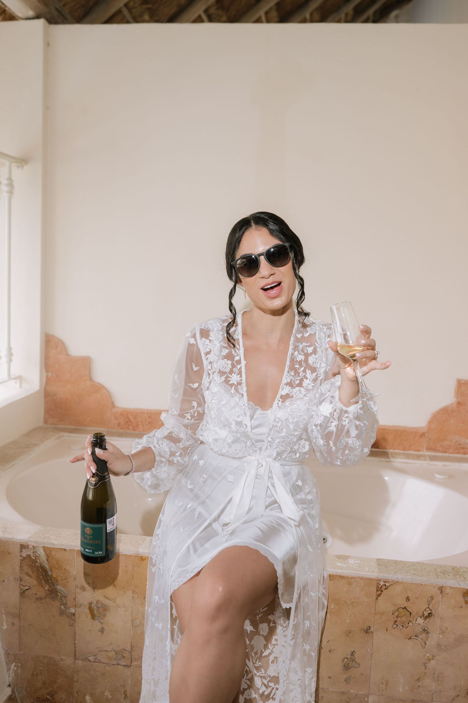 A person in a white lace robe poses with wine glasses in a rustic interior setting.