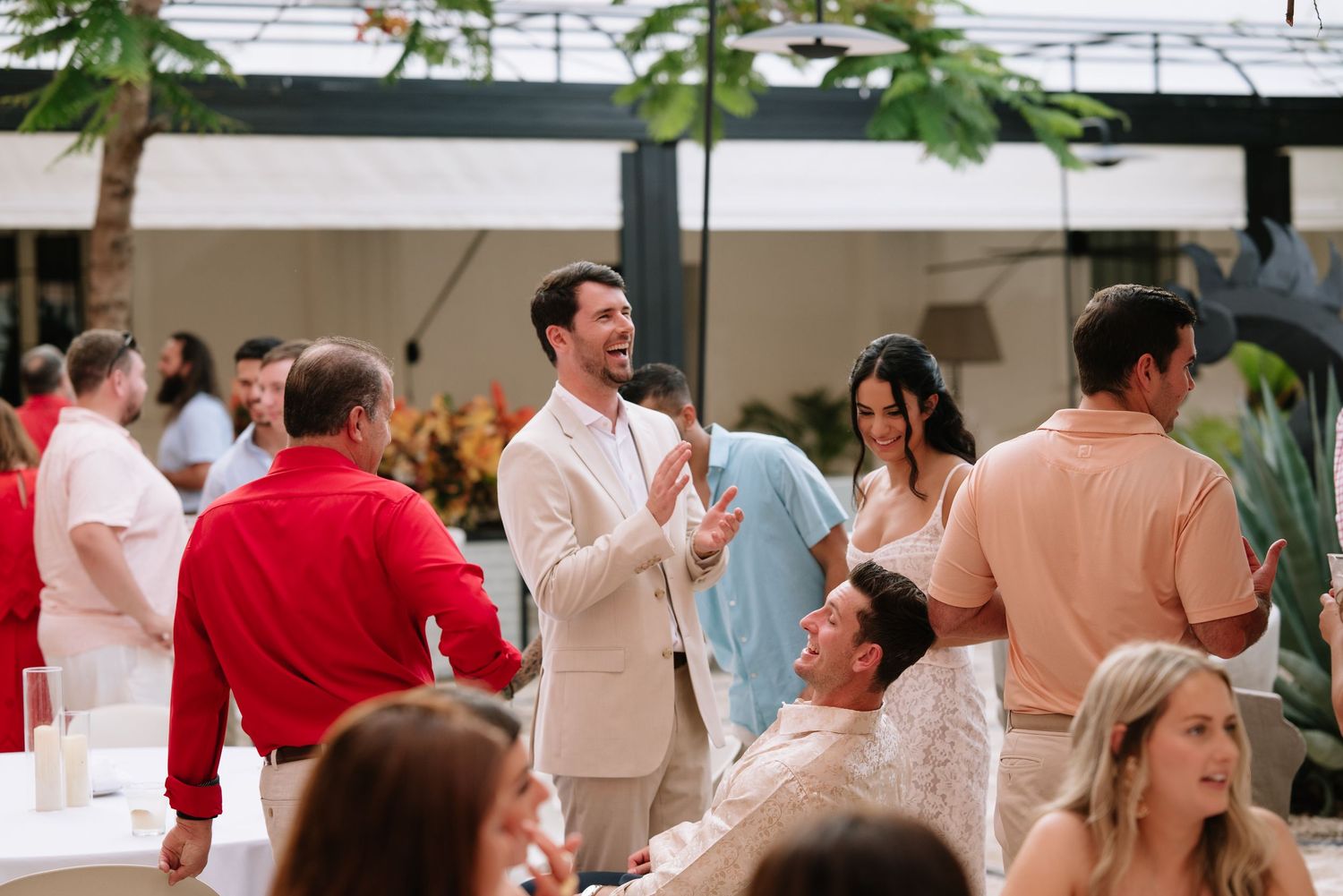 Wedding guests in light summer attire gather at an elegant indoor reception venue with tropical decor.