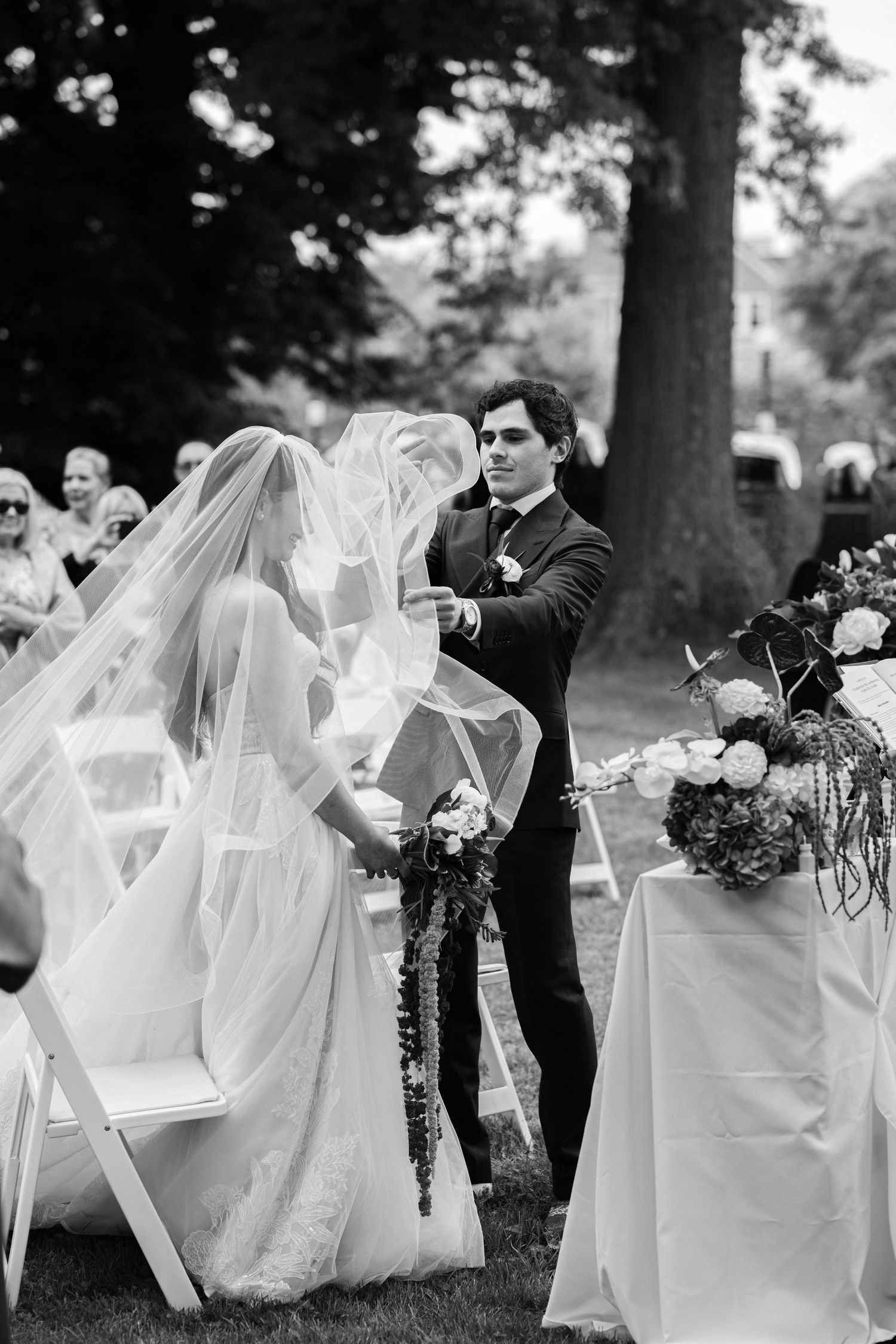 Groom lifts bride's veil during outdoor wedding ceremony, surrounded by floral arrangements and seated guests.