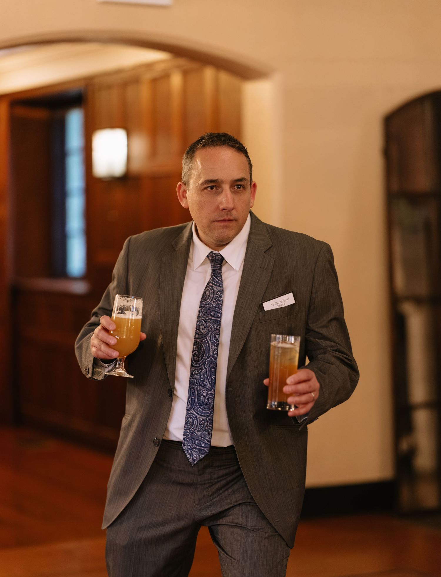 A man in a gray suit and paisley tie carries two glasses of beer at a formal wedding reception venue.