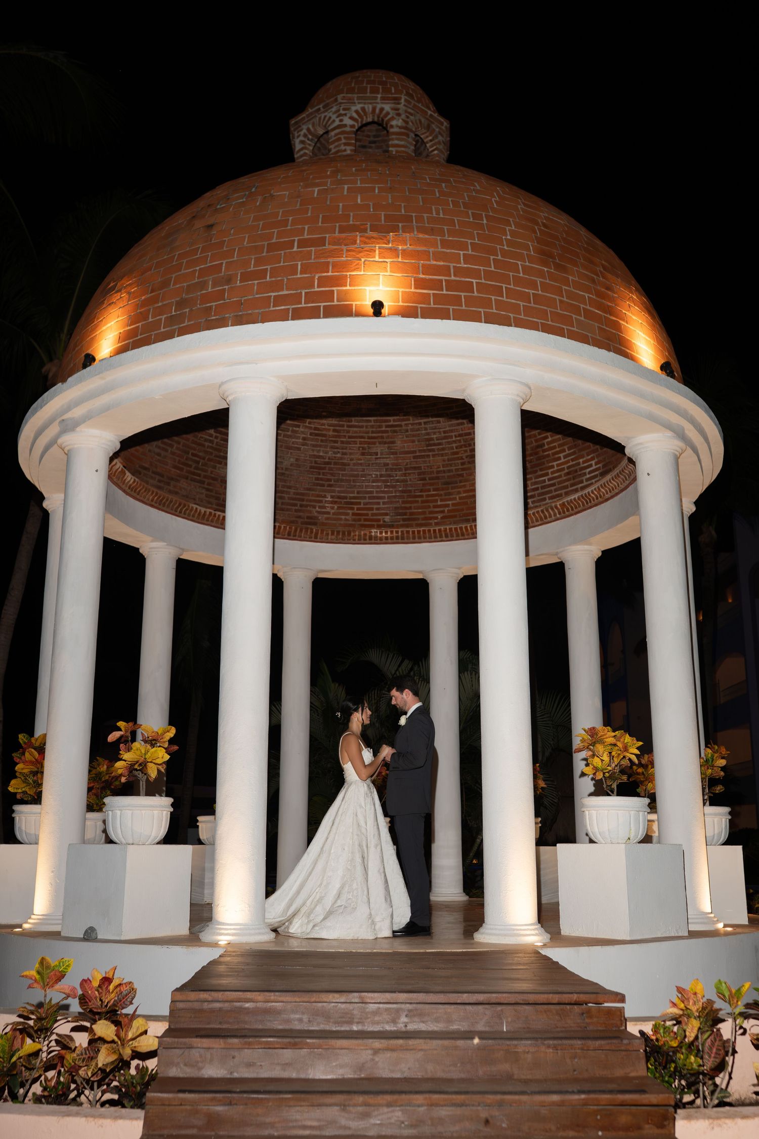 A romantic nighttime wedding scene under an illuminated white gazebo decorated with yellow flowers.