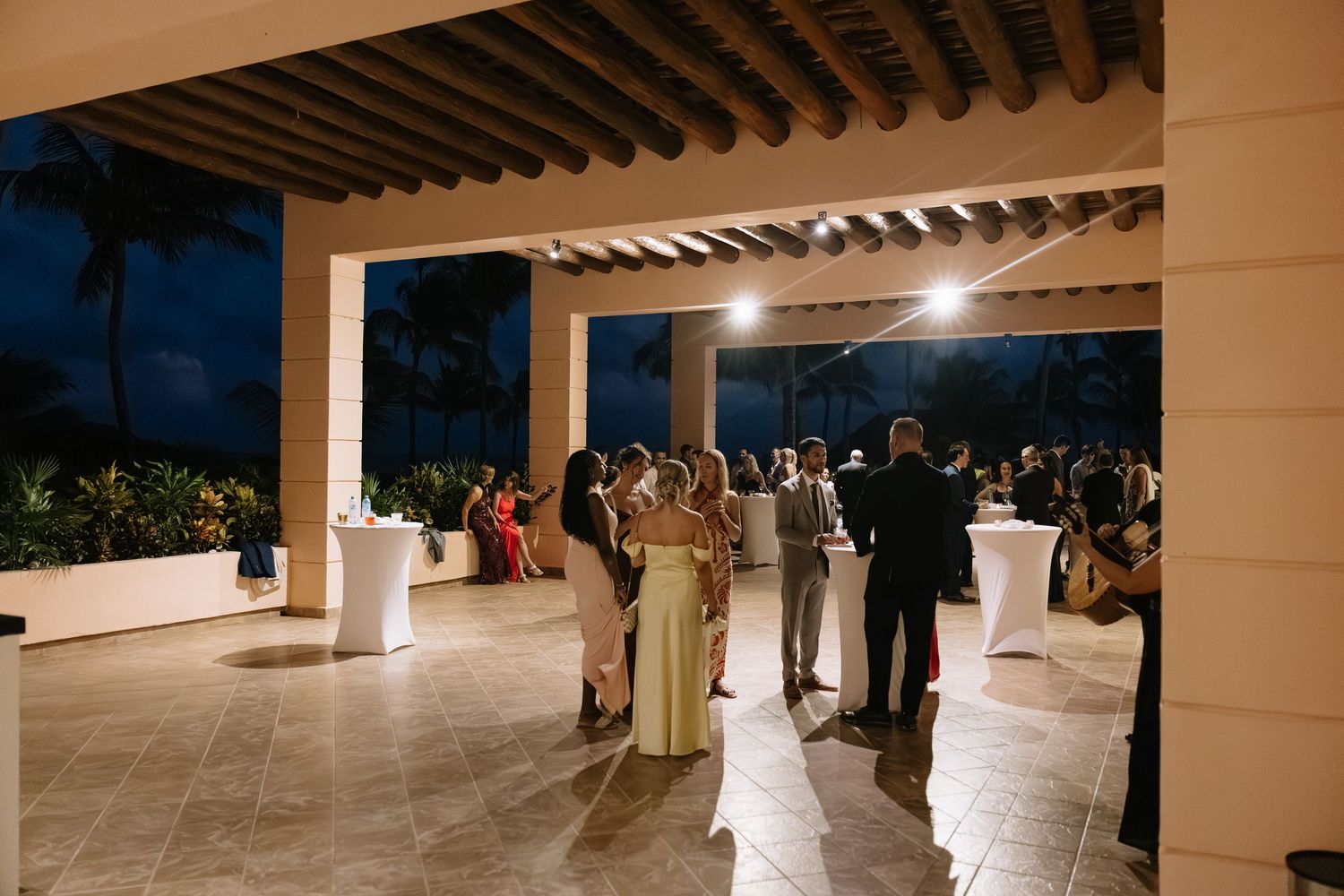 Evening wedding reception guests mingle on an elegant covered terrace with white columns and mood lighting.