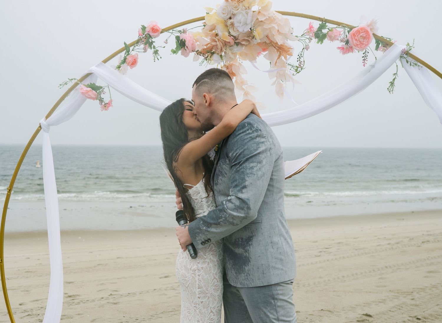A romantic couple shares an intimate moment under a white draped arch on a misty beach.