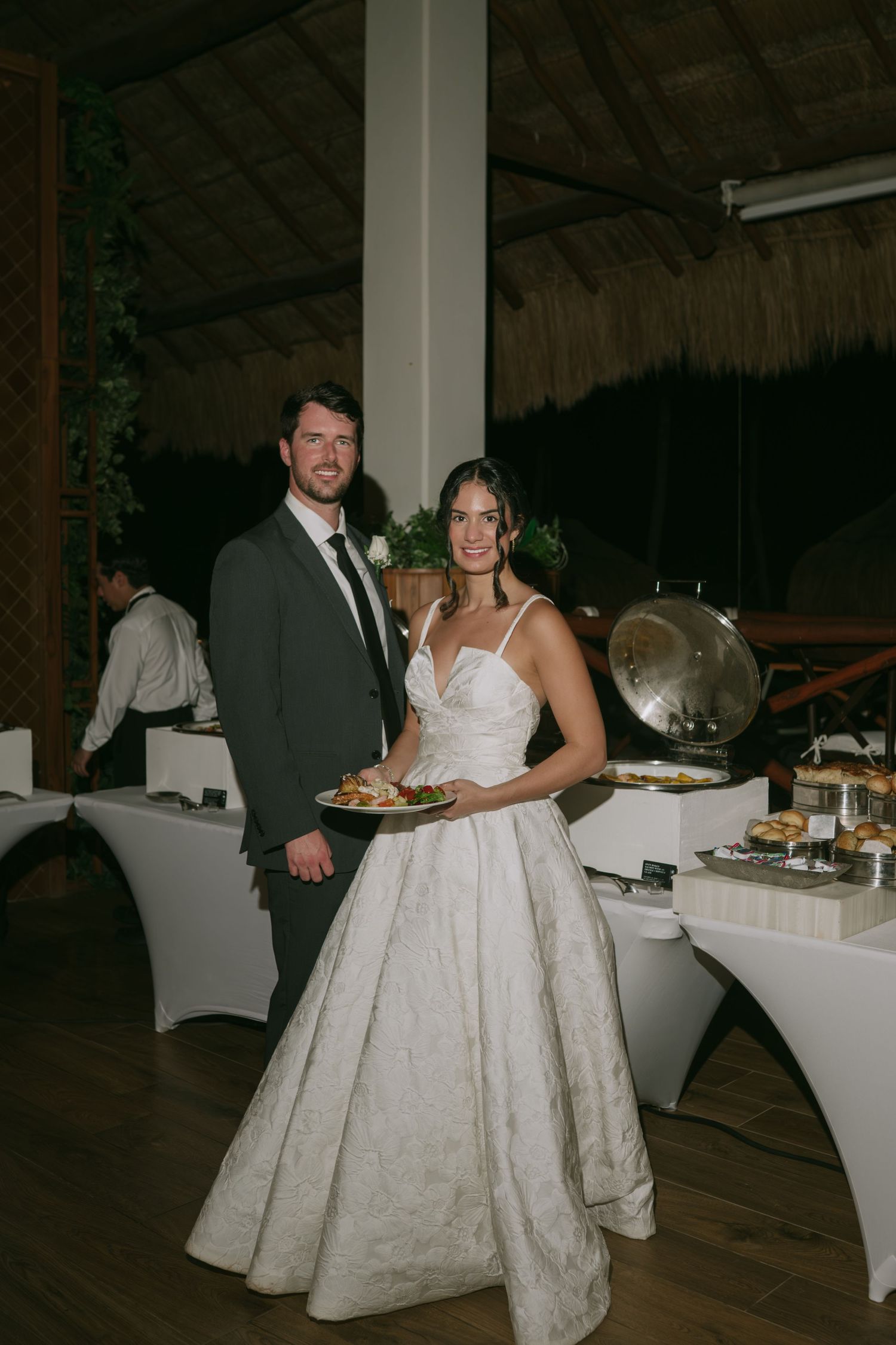 A couple at their wedding reception standing by the buffet table in an elegant venue with thatched roof decor.