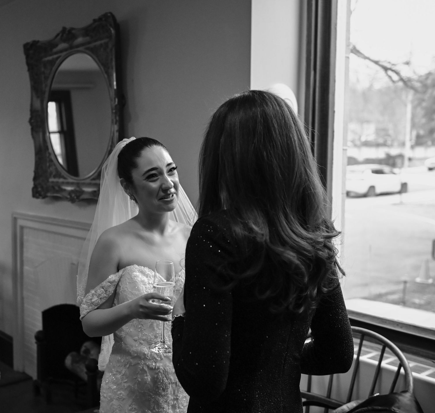 A bride in a white dress converses with a guest near an ornate mirror.