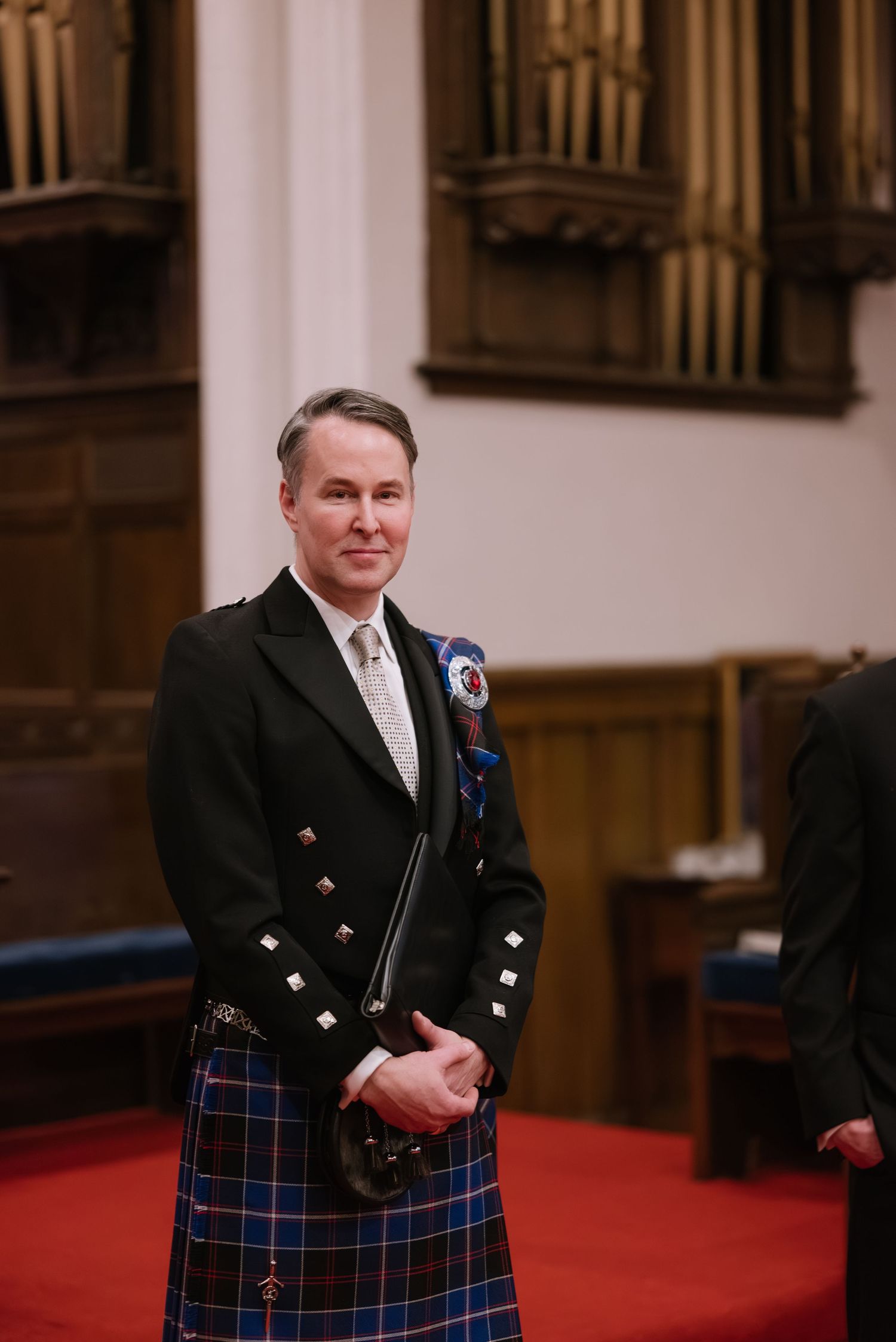 A man in traditional Scottish formal wear with kilt stands in a church interior.