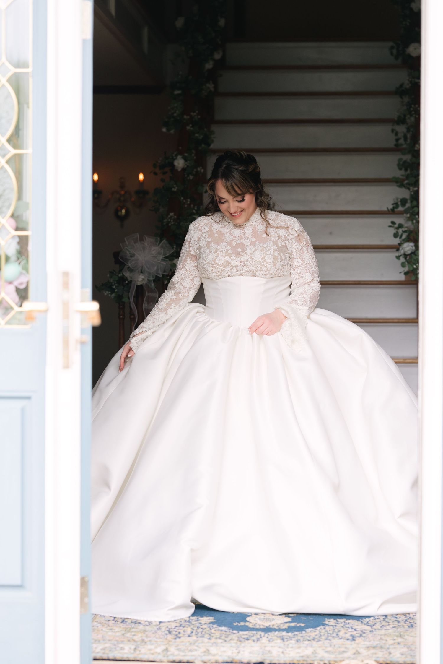 Bride in an elegant lace and tulle wedding gown sitting on white stairs with decorative railings.