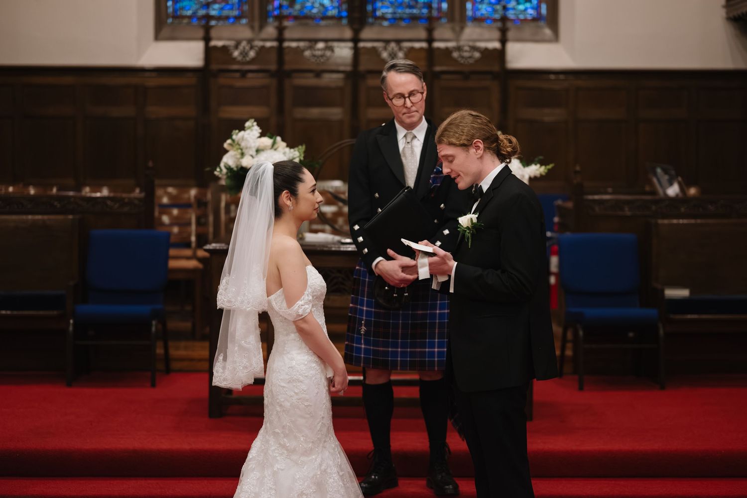 Wedding ceremony in progress at church altar with red carpet and stained glass windows.