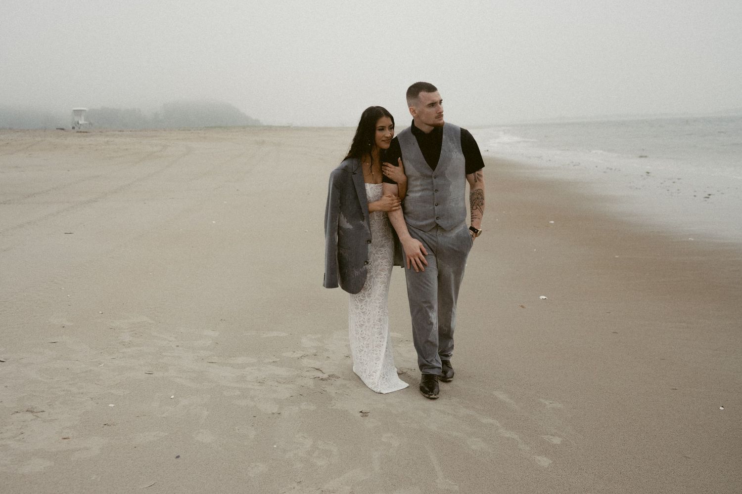 A couple walks together along a foggy beach shoreline during their wedding photoshoot.