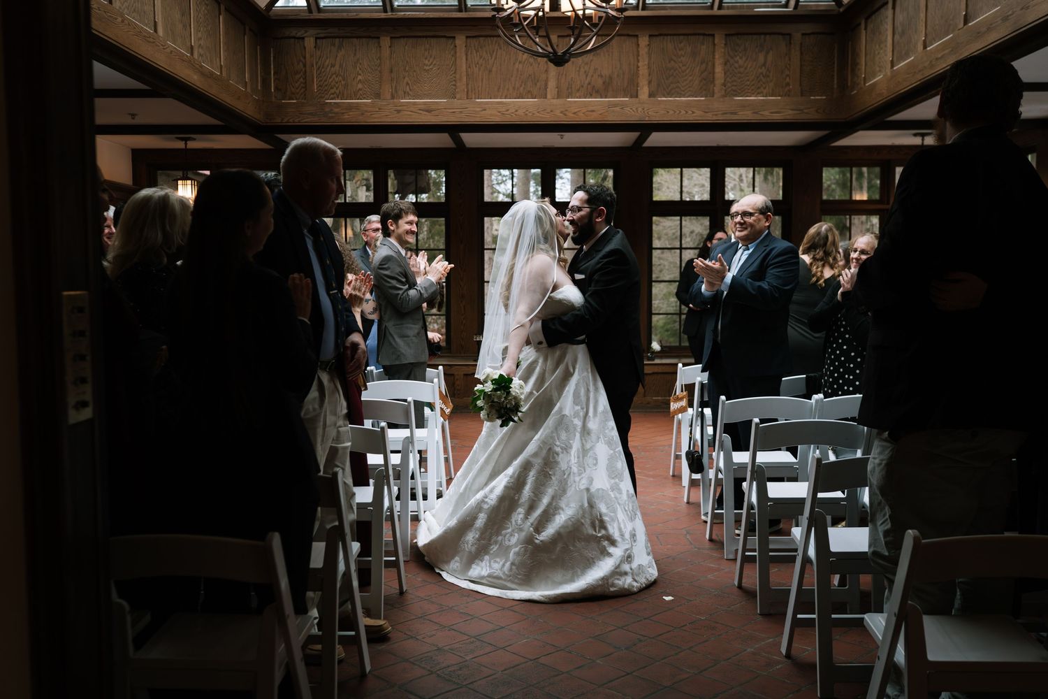 Bride and groom share a kiss at the altar during an elegant indoor wedding ceremony with white chairs and wood-paneled walls.