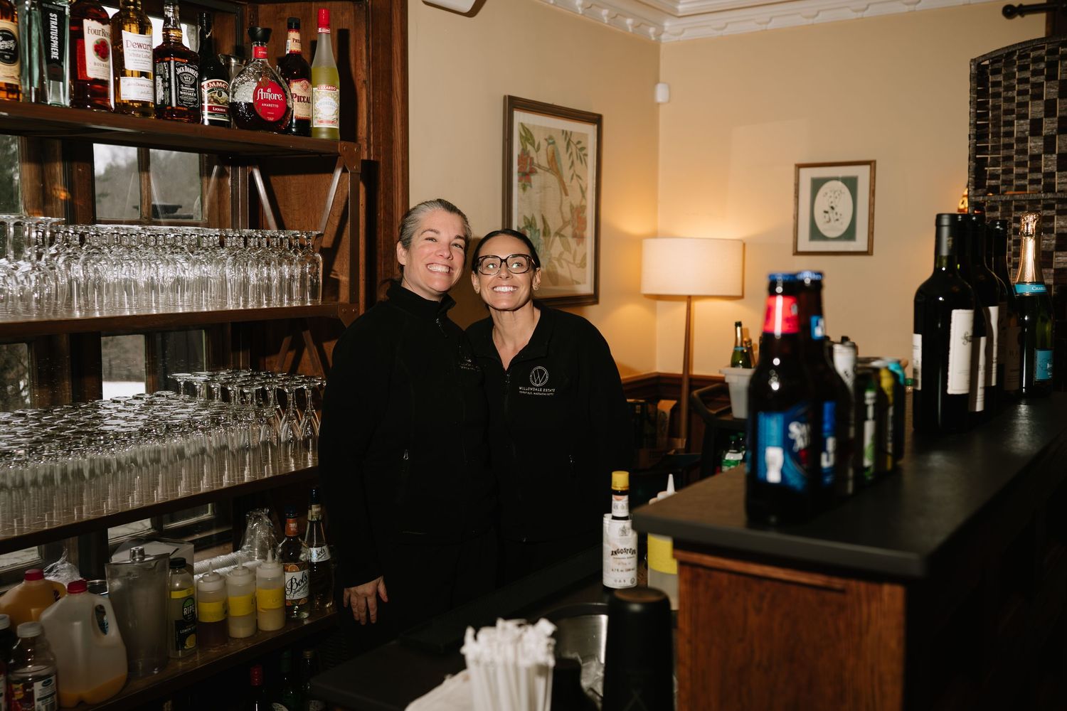 Two smiling bartenders in black uniforms pose behind a well-stocked bar with wine glasses and bottles.