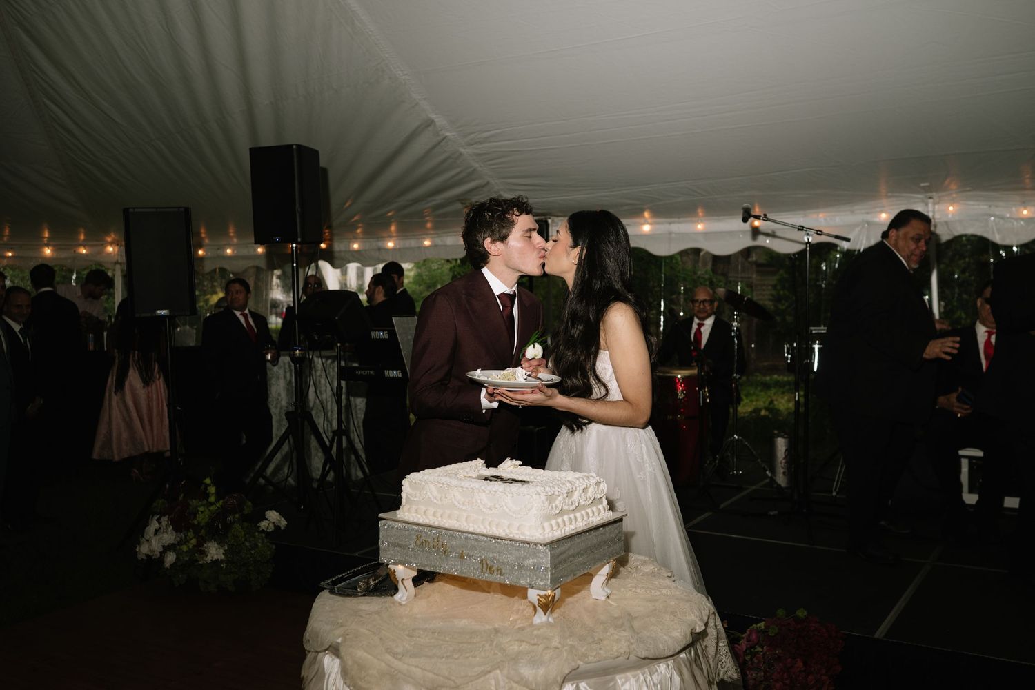 Newlyweds share a kiss while cutting their white wedding cake at a romantic tented reception celebration.