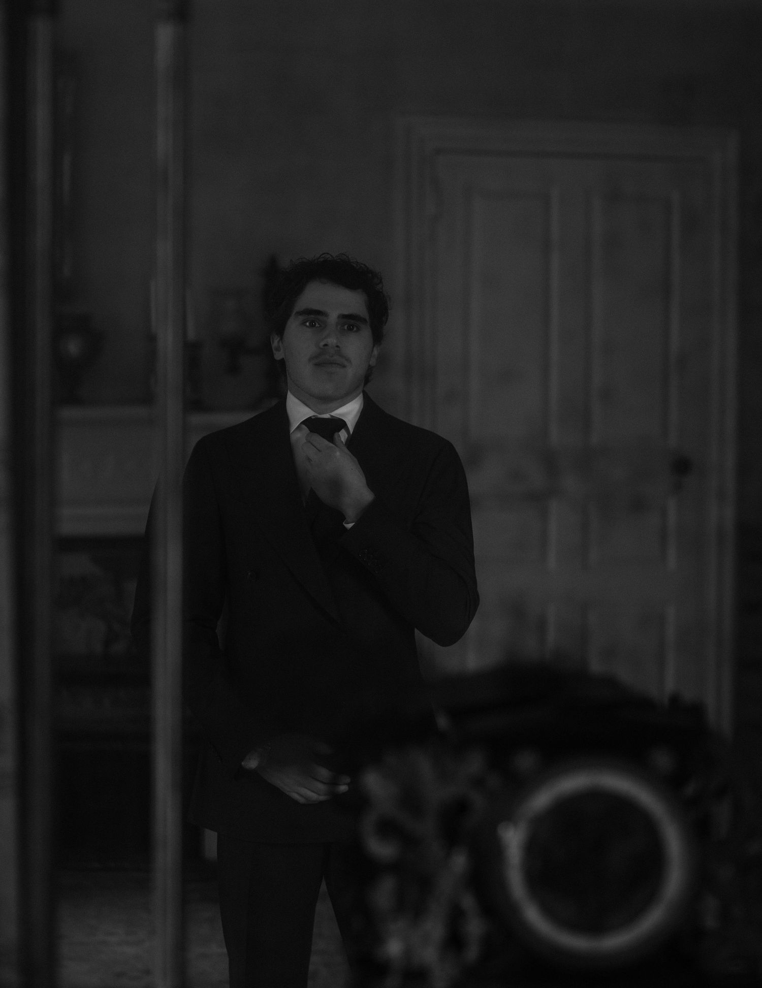 Black and white photo of a young man in a dark suit adjusting his tie indoors.