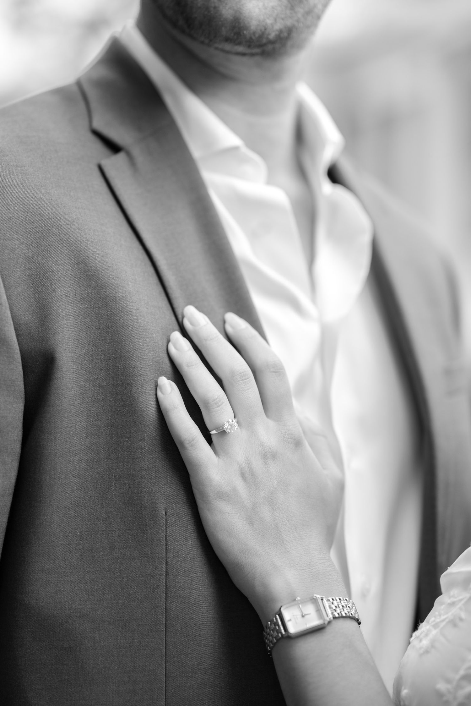 Close-up detail of hands with engagement ring against a gray suit jacket.