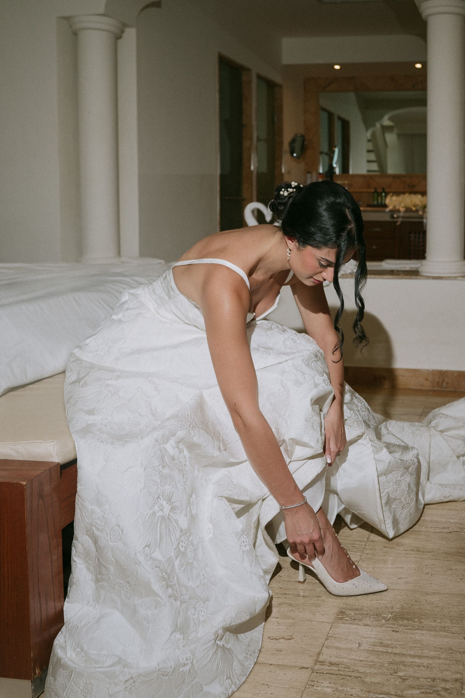 Bride in white wedding dress adjusting her elegant heels on a wooden floor.