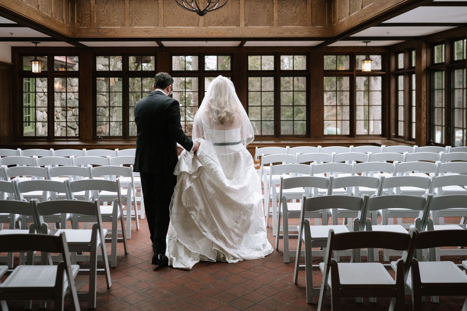 Bride and groom walk hand in hand through elegant wedding venue with white chairs and large windows.