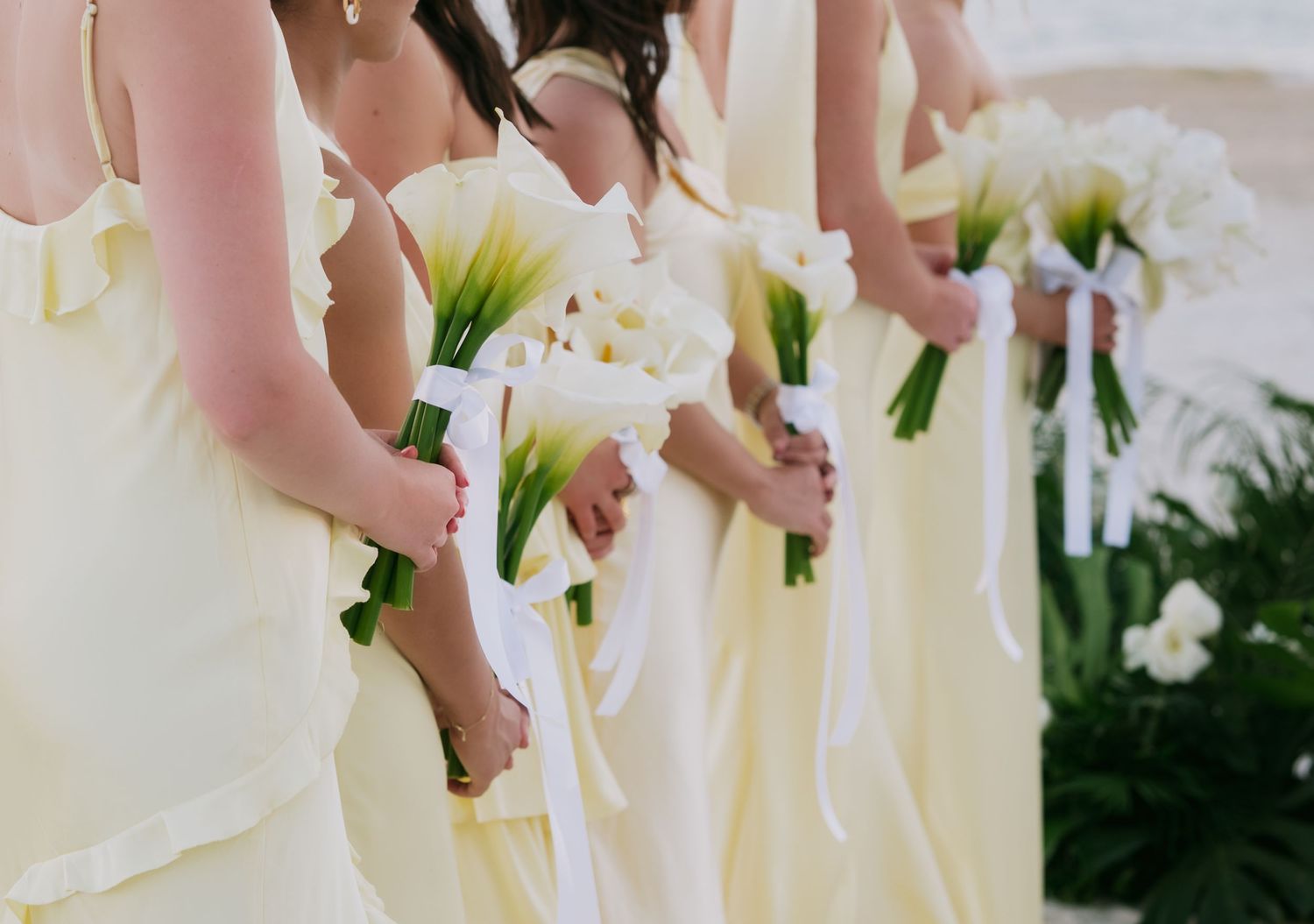 A row of bridesmaids in pale yellow dresses hold white floral bouquets against a garden backdrop.