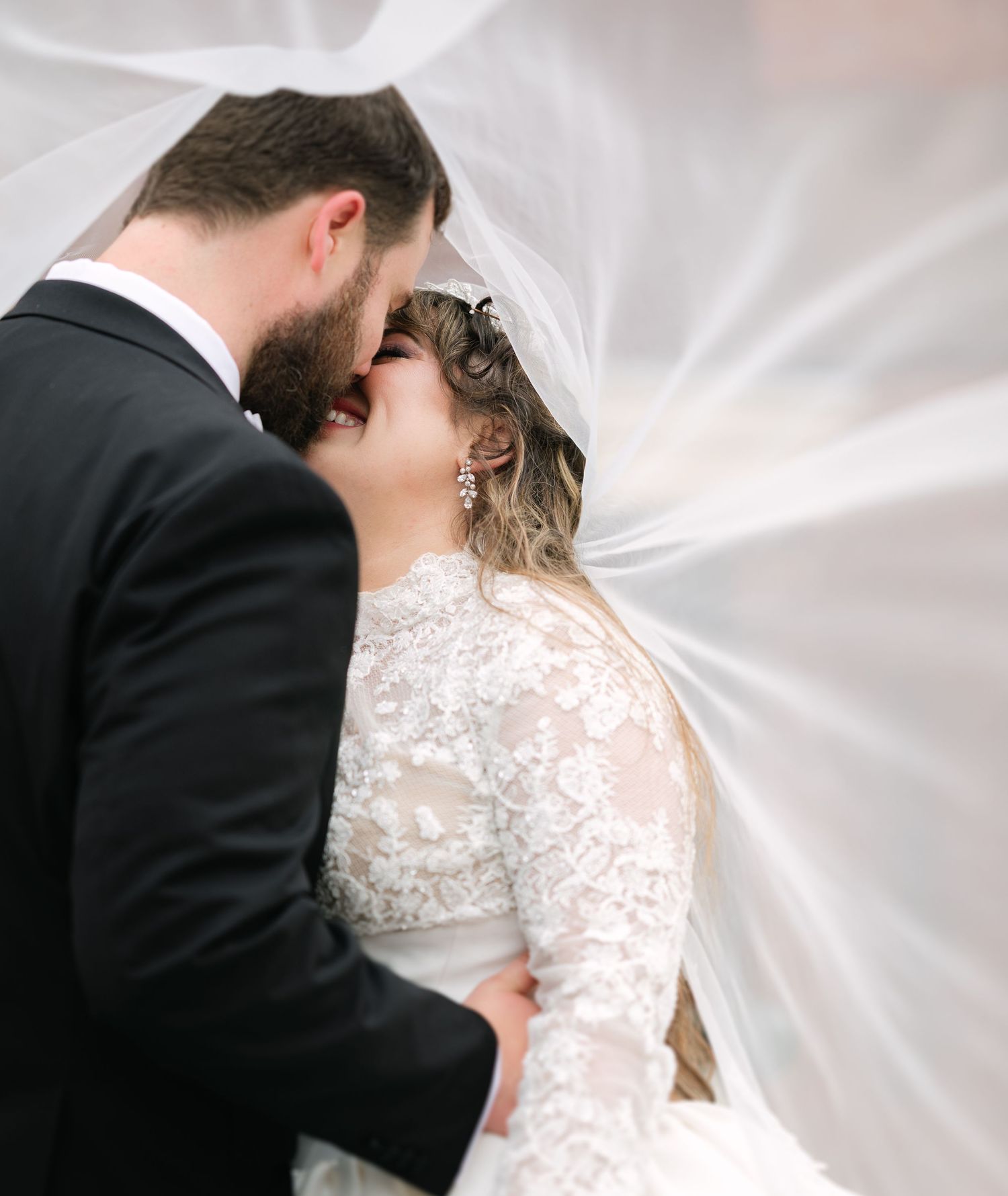 Romantic wedding couple embracing under a flowing white veil in soft light.