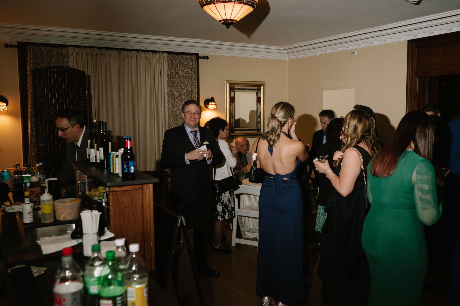Elegantly dressed guests mingle at a formal cocktail party near a well-stocked bar in a warmly lit venue.