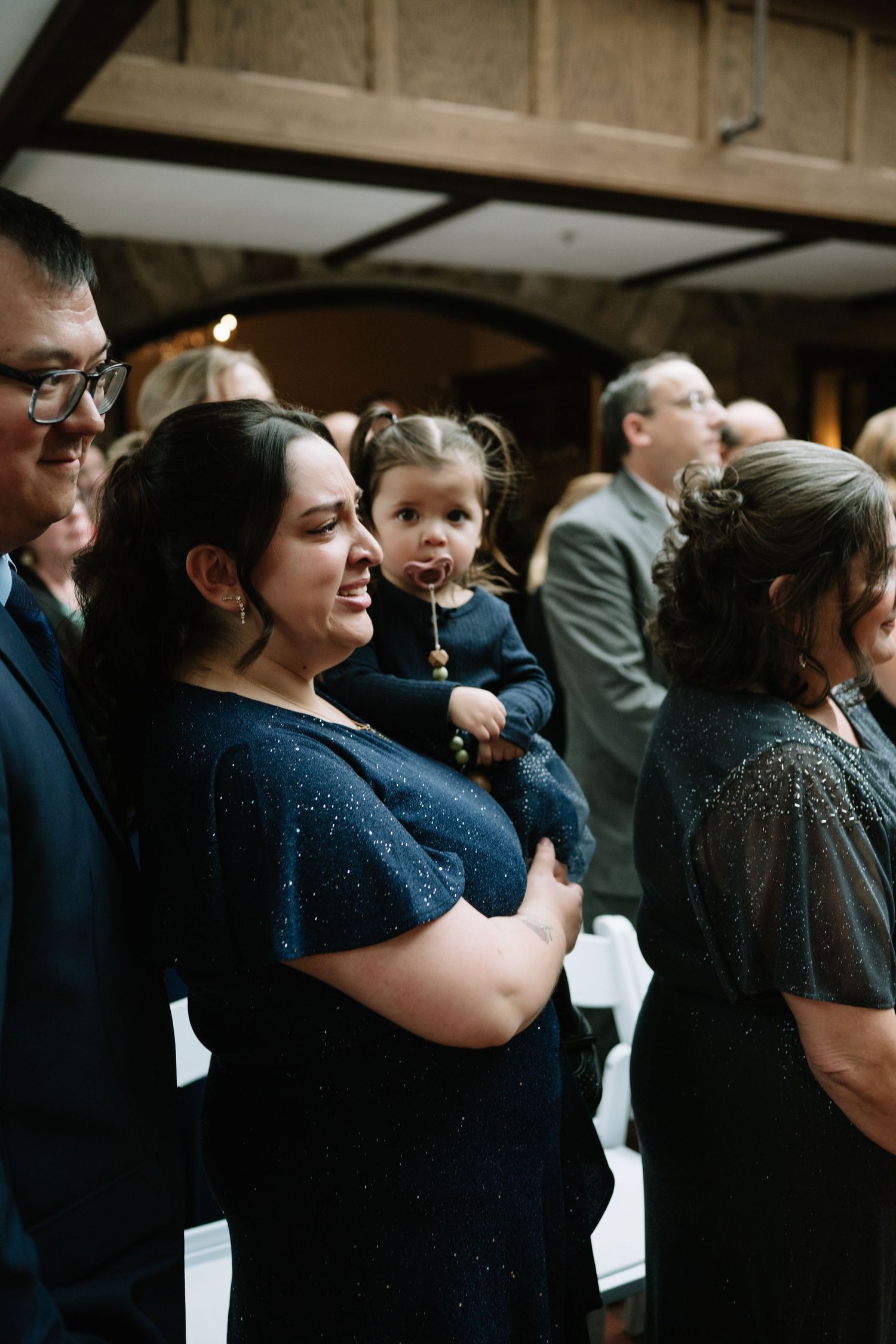 Guests in formal attire watch a wedding ceremony, woman in navy dress holds a toddler girl.